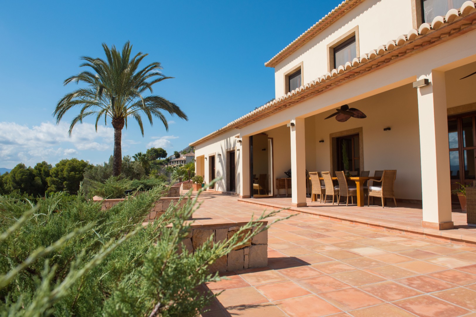 Terracotta-Terrasse mit überdachtem Essbereich und Korbstühlen, umgeben von einer Palme und mediterraner Vegetation unter blauem Himmel.