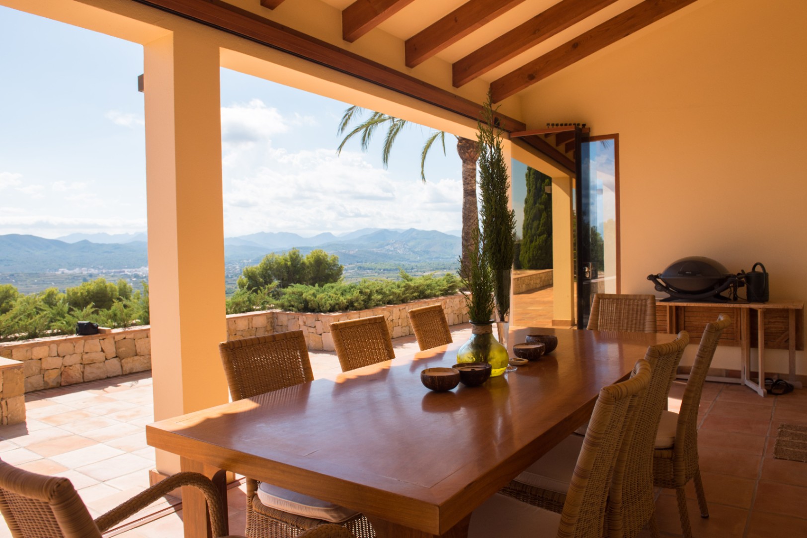 Überdachte Terrasse mit großem Holztisch und Rattanstühlen unter einer Sichtbalkendecke. Panoramablick auf die Berge und ein Außenbereich mit Natursteinmauer.