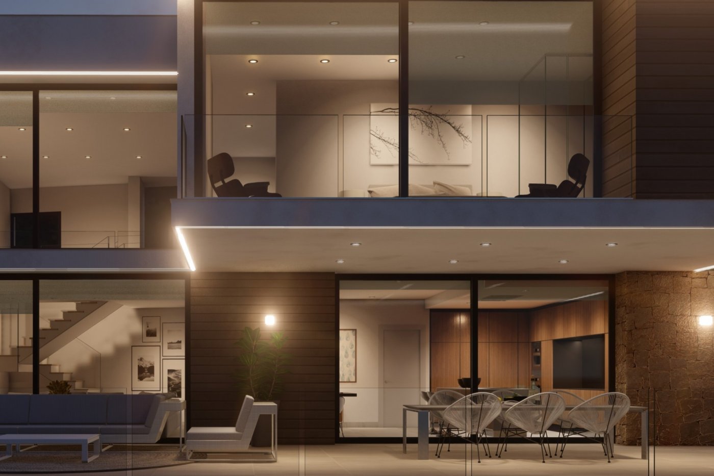 Außenansicht einer Villa bei Nacht mit Terrasse, Acapulco-Stühlen und Natursteinwänden. Große Glasfronten beleuchten die Küche und den oberen Balkon mit Glasgeländer.