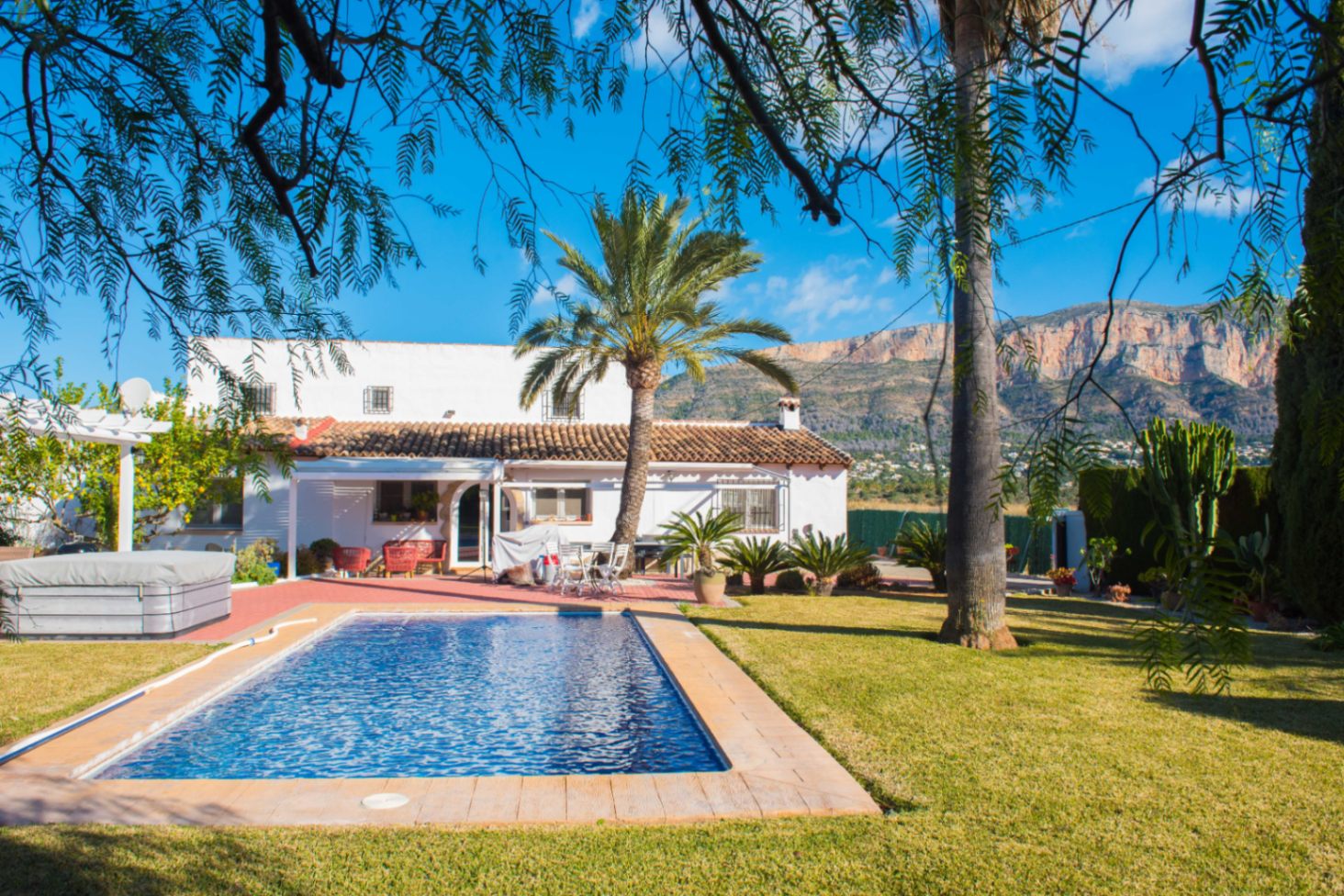Außenanlage mit rechteckigem Pool, Rasenfläche und Palmen vor dem Montgó-Massiv. Die weiße Villa verfügt über ein Terrakottadach und einen Whirlpool im Terrassenbereich.