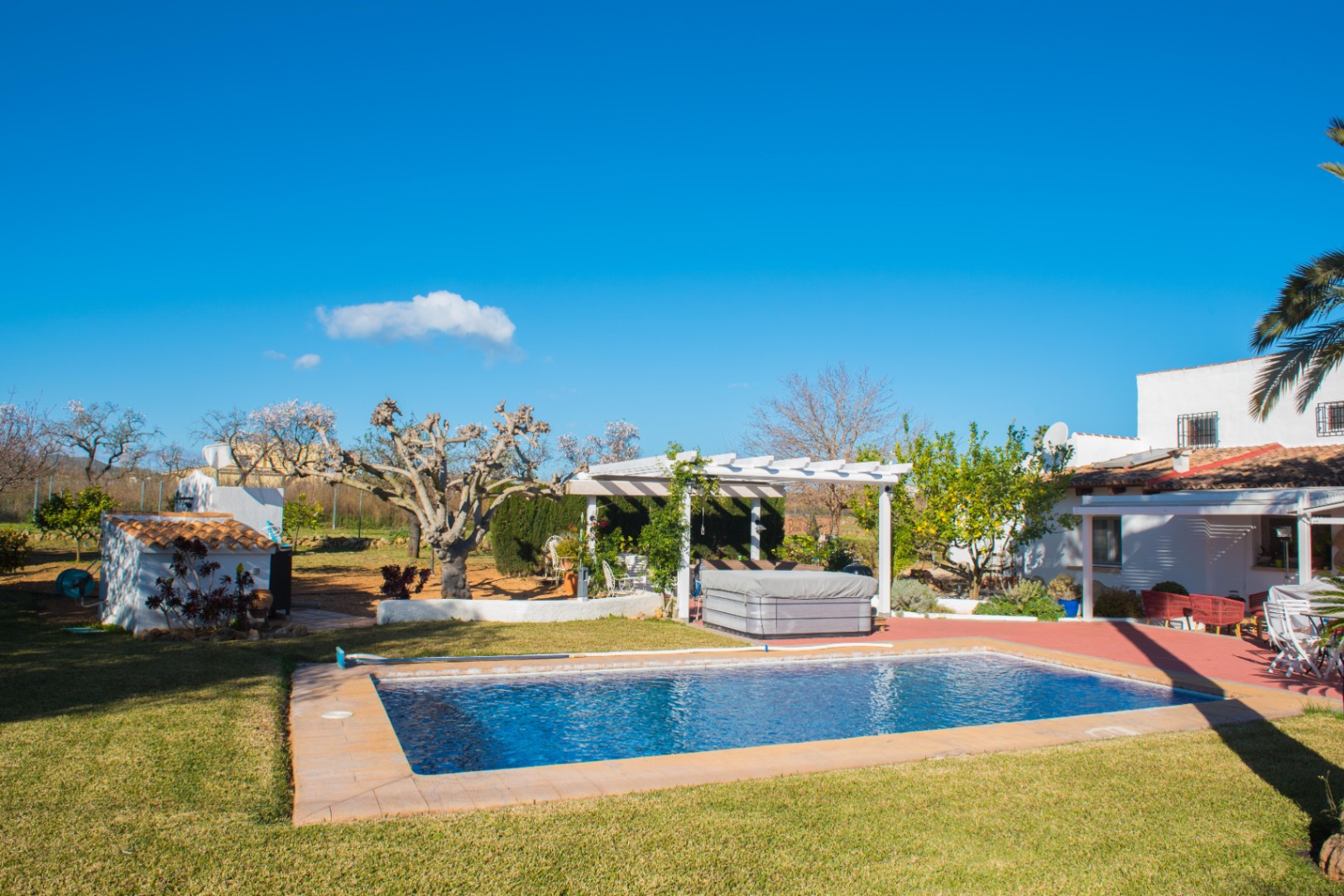 Außenbereich mit rechteckigem Pool, gepflegtem Rasen und einer weißen Pergola mit Whirlpool, umgeben von einem mediterranen Garten und Wohnhaus.
