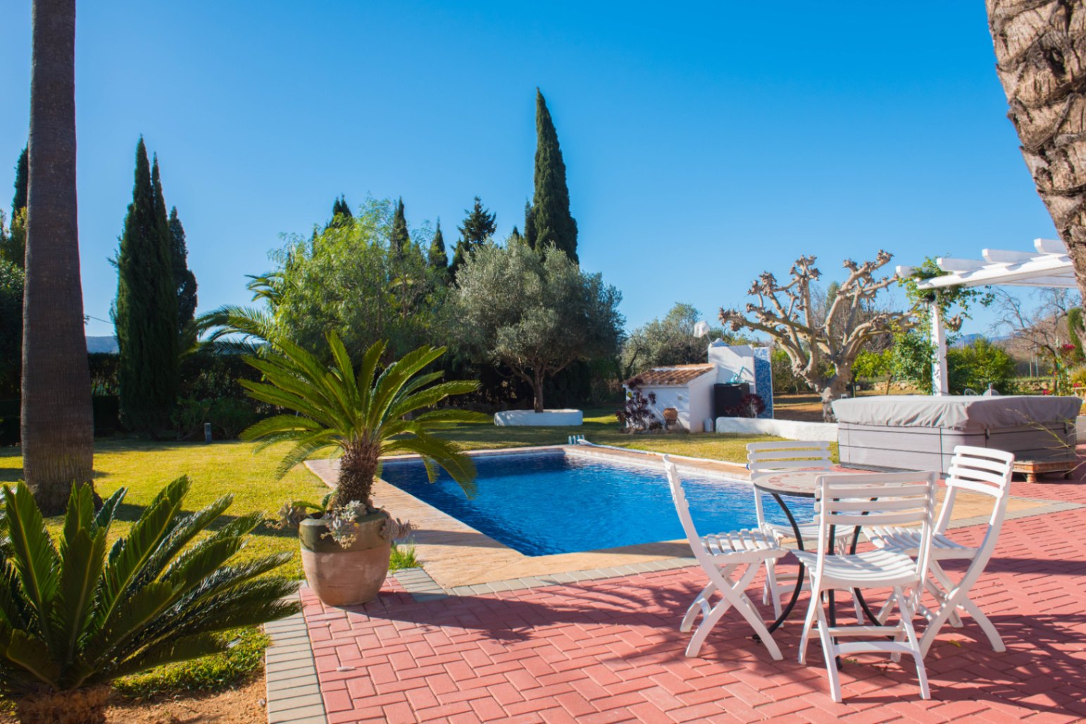 Außenbereich mit rotem Backsteinpflaster, rechteckigem Pool, weißen Gartenmöbeln und einer gepflegten Rasenfläche mit mediterraner Bepflanzung.
