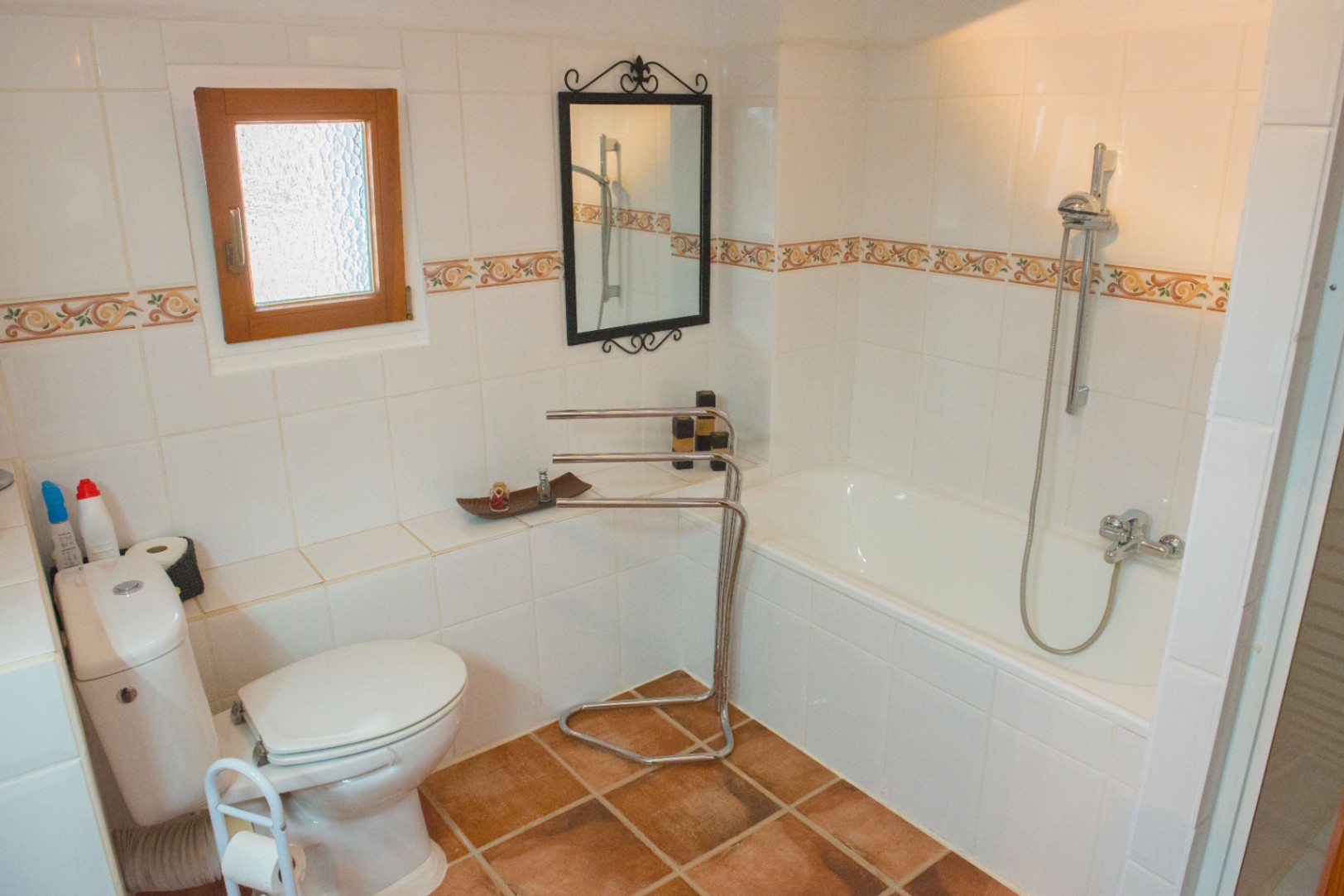 Badezimmer mit Einbaubadewanne, Terrakottaboden und weißen Wandfliesen mit Zierbordüre. Ausgestattet mit einem Holzfenster und einem Schmiedeeisenspiegel.