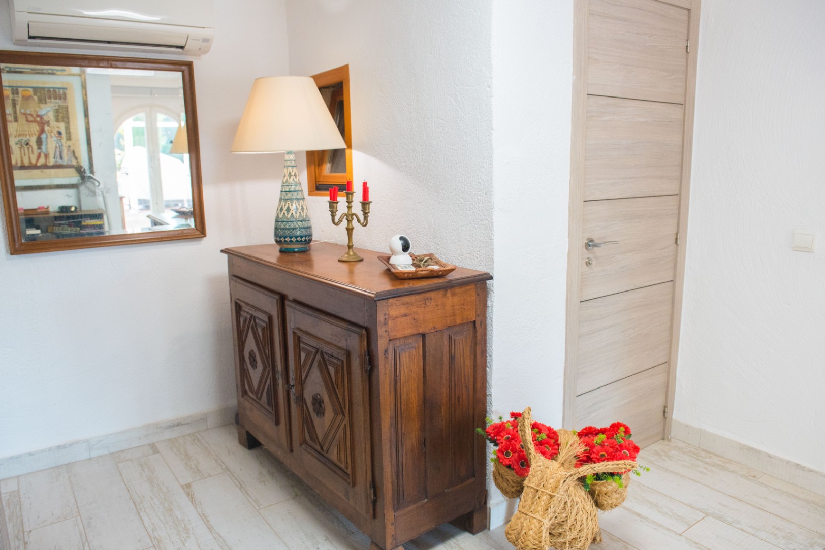 Eingangsbereich mit geschnitztem Sideboard aus dunklem Holz, Keramiktischlampe und hellem Boden in Holzoptik. Spiegel und Klimaanlage an der weiß verputzten Wand.