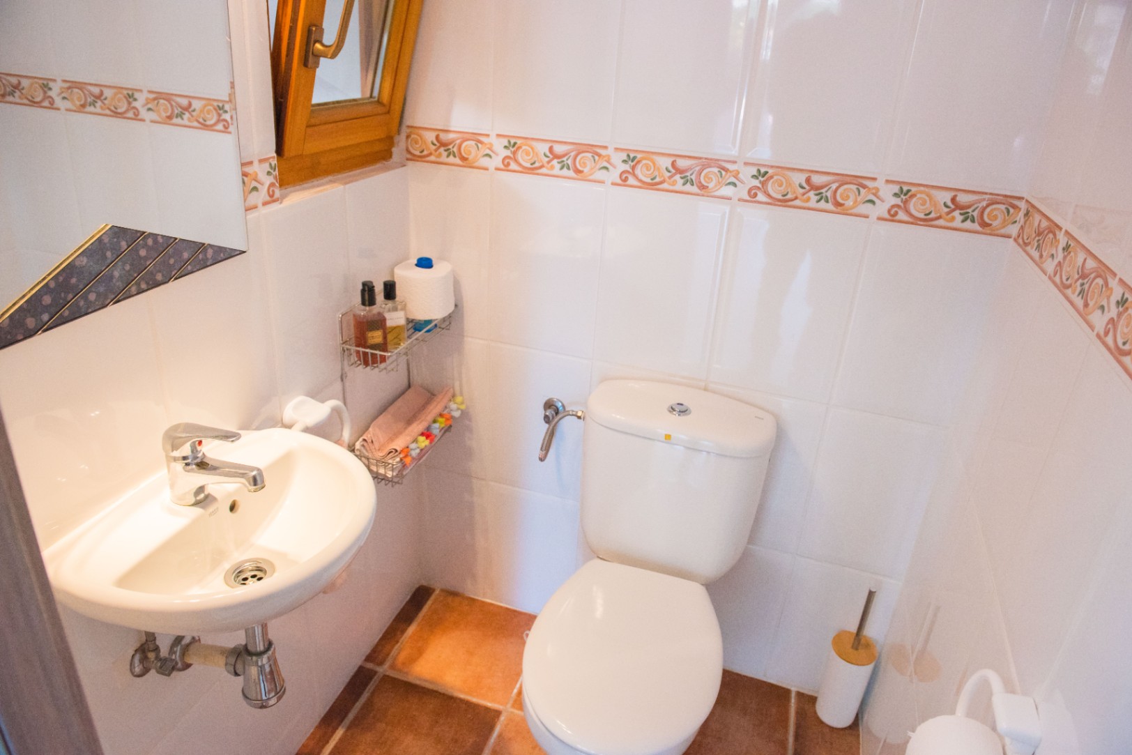 Kompaktes Gäste-WC mit weißen Wandfliesen und dekorativer Bordüre, Terrakottaboden, wandmontiertem Waschbecken und Holzfenster zur natürlichen Belüftung.