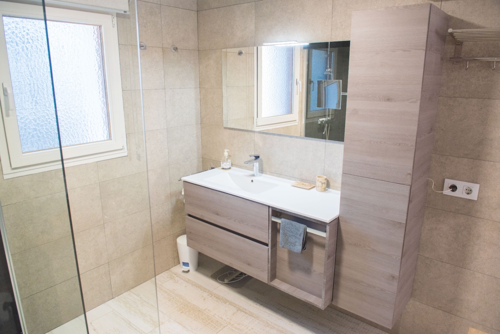 Modernes Badezimmer mit schwebendem Waschtisch in Holzoptik, passendem Hochschrank und großformatigen sandfarbenen Wandfliesen.
