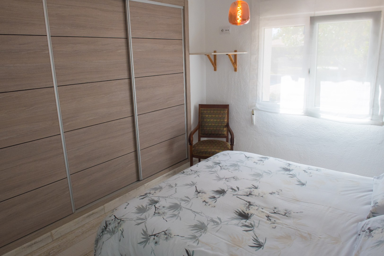 Schlafzimmer mit einem großen Einbauschrank mit Schiebetüren in Holzoptik, einem klassischen Sessel und natürlichem Licht durch das Fenster.