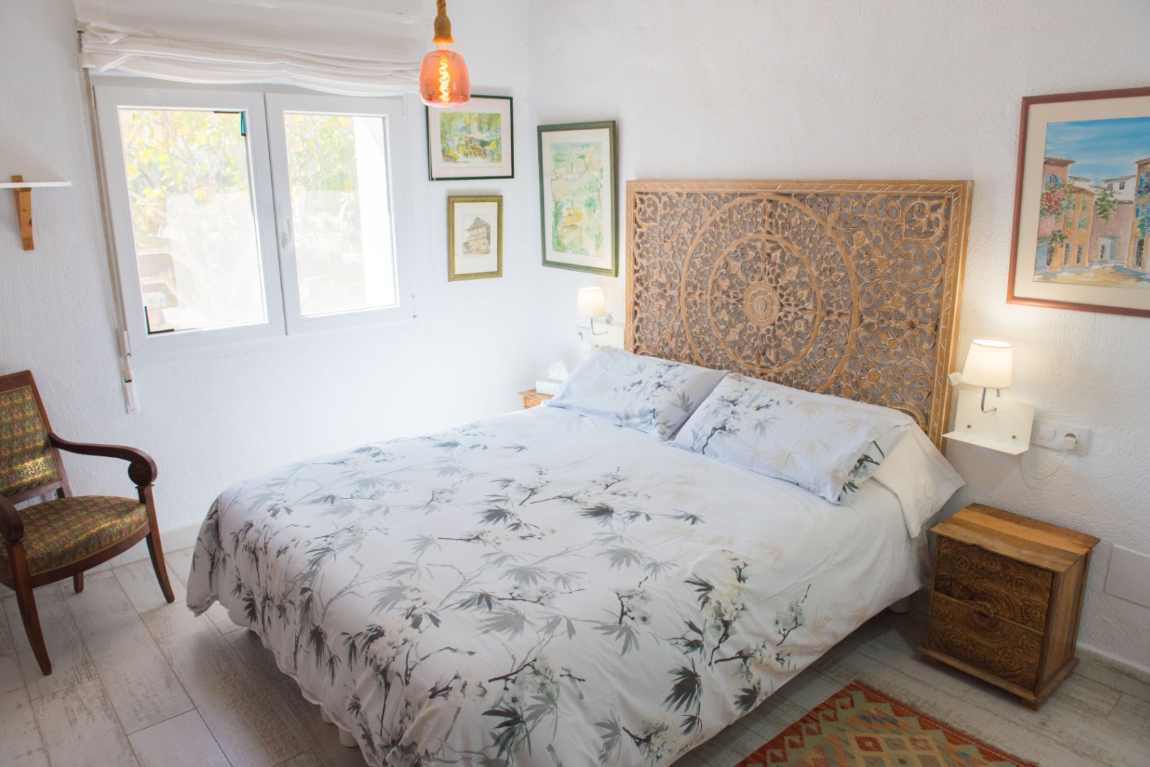 Schlafzimmer mit geschnitztem Holzkopfteil, floraler Bettwäsche und Vintage-Sessel. Weiße Wände mit Kunstwerken und Bodenbelag in heller Holzoptik.