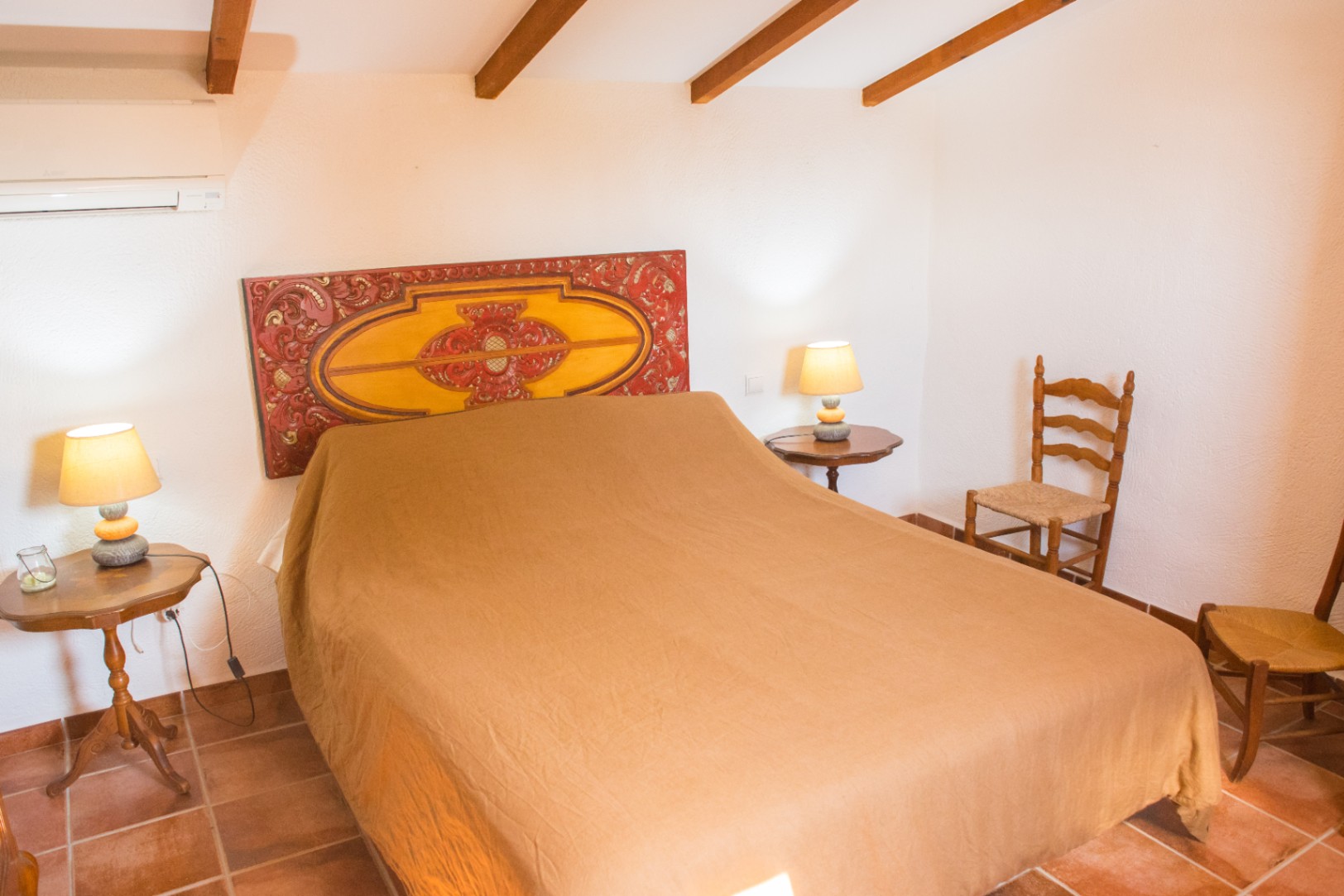 Schlafzimmer mit kunstvoll geschnitztem Holzkopfteil, sichtbaren Deckenbalken, Terrakottafliesen und rustikalen Holzmöbeln.