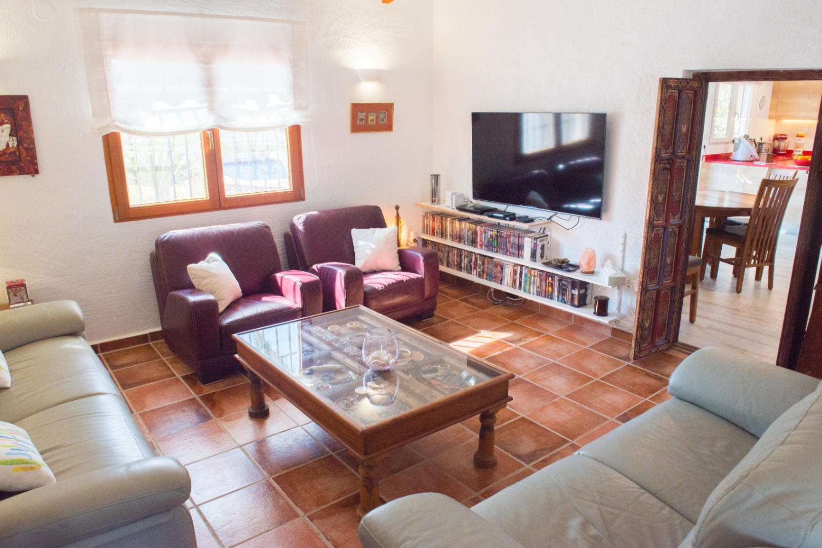 Wohnzimmer mit Terrakotta-Fliesenboden, Ledersofas und Glastisch. Wandmontierter Fernseher mit Medienregalen neben einem kunstvoll geschnitzten Holztürrahmen.