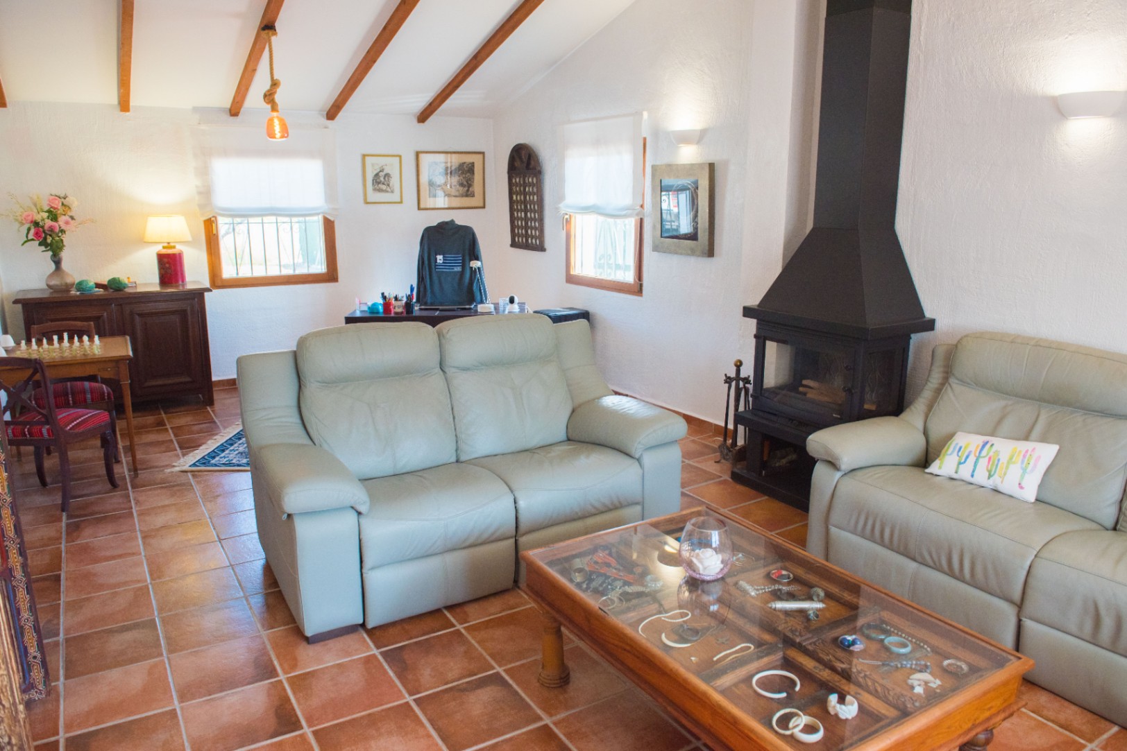 Wohnzimmer mit Terrakotta-Fliesenboden, schwarzem Metallkamin und Dachschräge mit sichtbaren Holzbalken. Ausgestattet mit Ledersofas und einem Couchtisch mit Glasplatte.