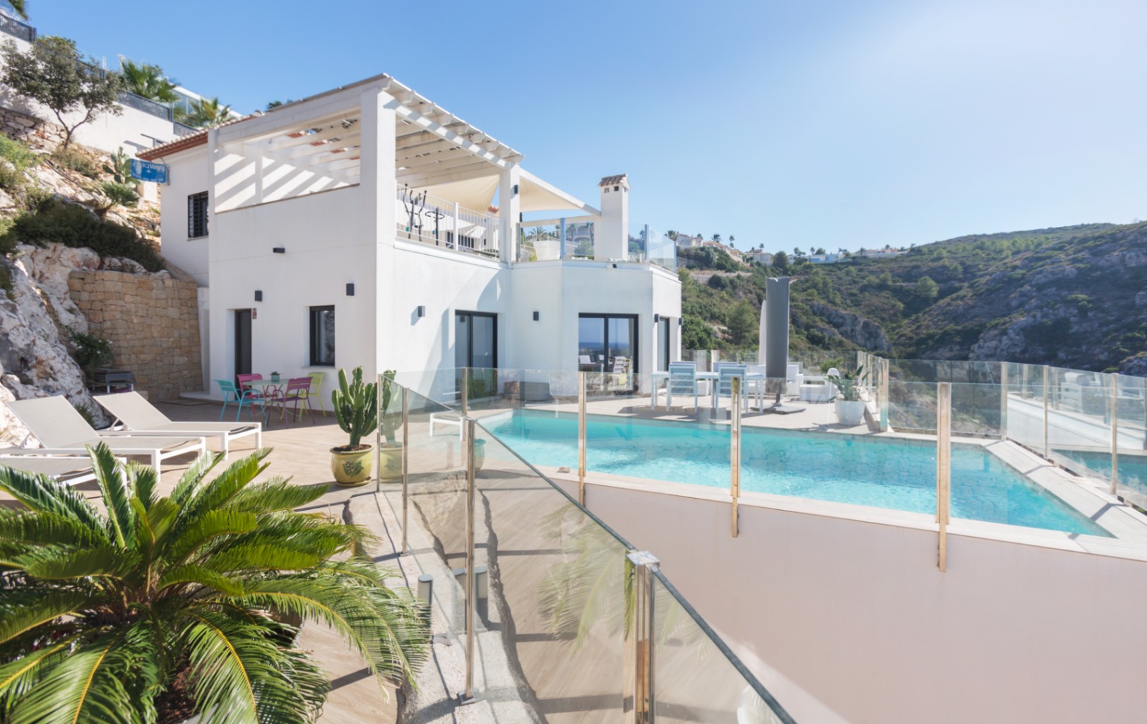 Außenansicht einer weißen, mehrstöckigen Villa mit Pool, Glasgeländern, Sonnenterrassen und Panoramablick auf die umliegenden Hügel.