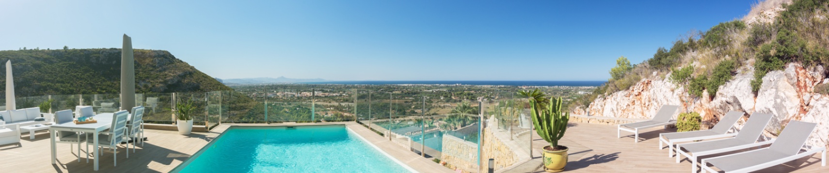 Erhöhte Terrasse mit Pool, Glasgeländern und weißen Außenmöbeln sowie Panoramablick auf das Tal und die Mittelmeerküste.