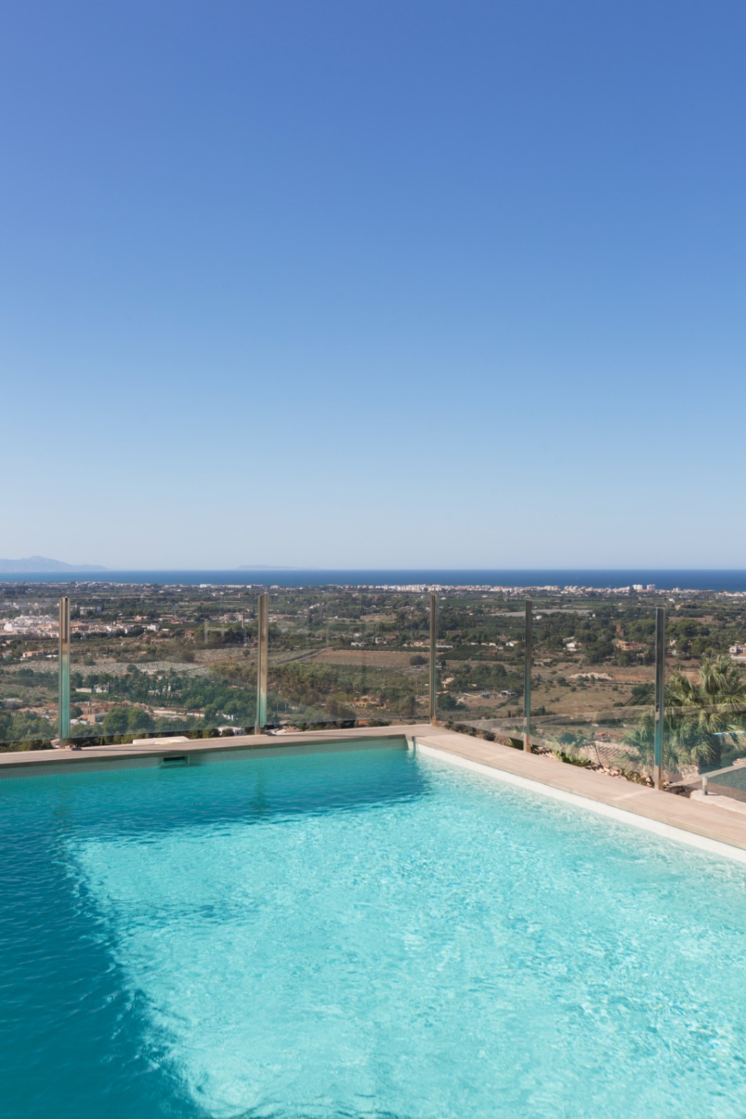 Erhöhter Infinity-Pool mit Glasgeländer und Panoramablick über das Küstental bis hin zum Mittelmeer unter klarem blauem Himmel.