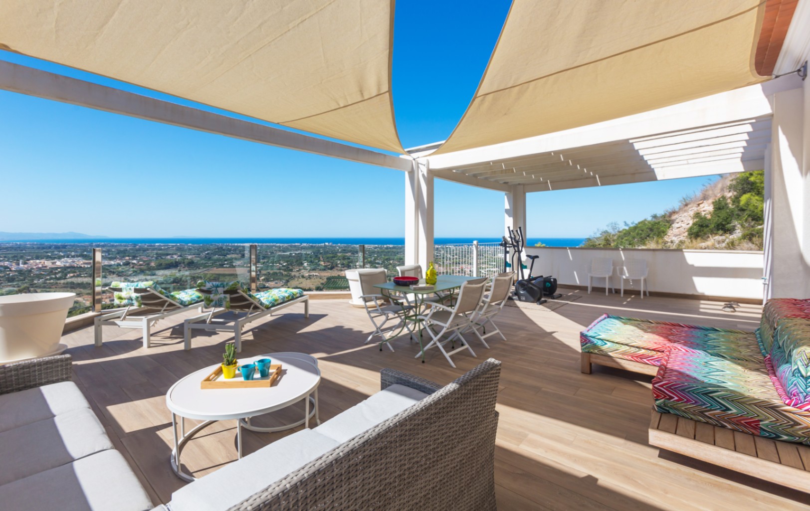 Große Dachterrasse mit Bodenfliesen in Holzoptik, Sonnensegeln und Panoramablick auf das Meer. Ausgestattet mit Essbereich, Lounge-Möbeln und Sonnenliegen unter einer Pergola.