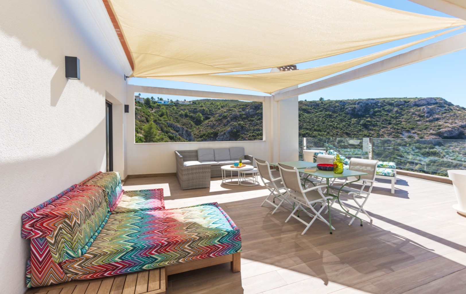 Großzügige Terrasse mit beigen Sonnensegeln, Essgruppe, Korb-Lounge und buntem Daybed mit Blick auf das grüne Tal durch Glasgeländer.