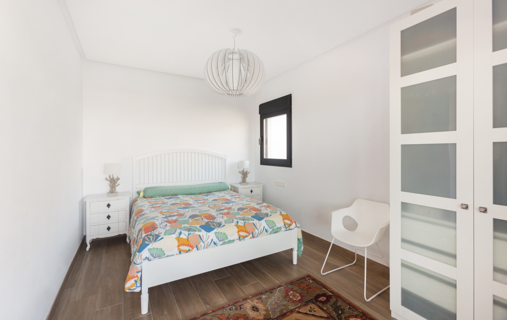 Helles Schlafzimmer mit weißem Holzbett, passenden Nachttischen und einem Kleiderschrank mit Milchglaseinsätzen. Boden in Holzoptik und eine moderne Designer-Pendelleuchte.