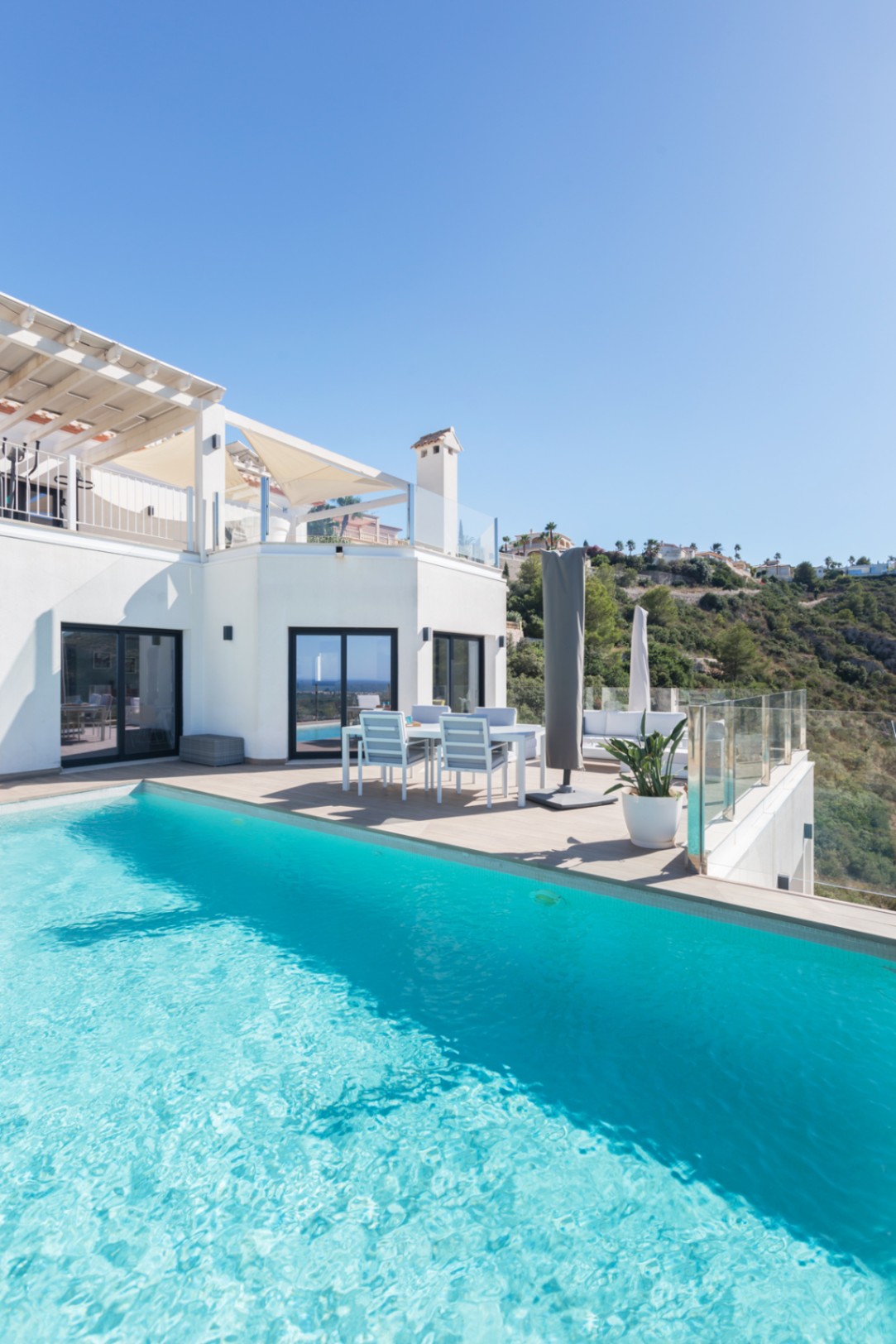 Infinity-Pool mit türkisfarbenem Wasser neben einer Terrasse mit Essbereich, Glasgeländern und Panoramablick auf die umliegende Hügellandschaft.