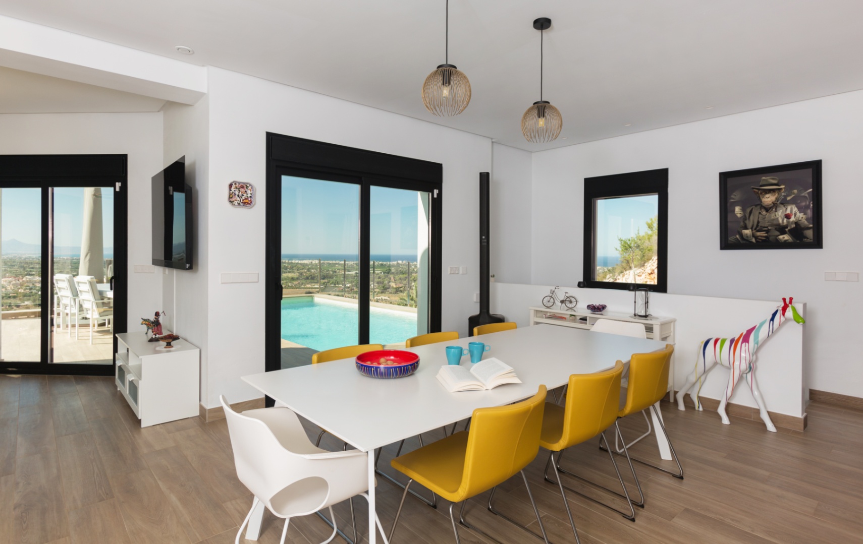 Moderner Essbereich mit weißem Tisch, gelben Stühlen und Holzeffekt-Fliesen, der sich durch Glasschiebetüren zur Poolterrasse mit Weitblick auf das Meer öffnet.