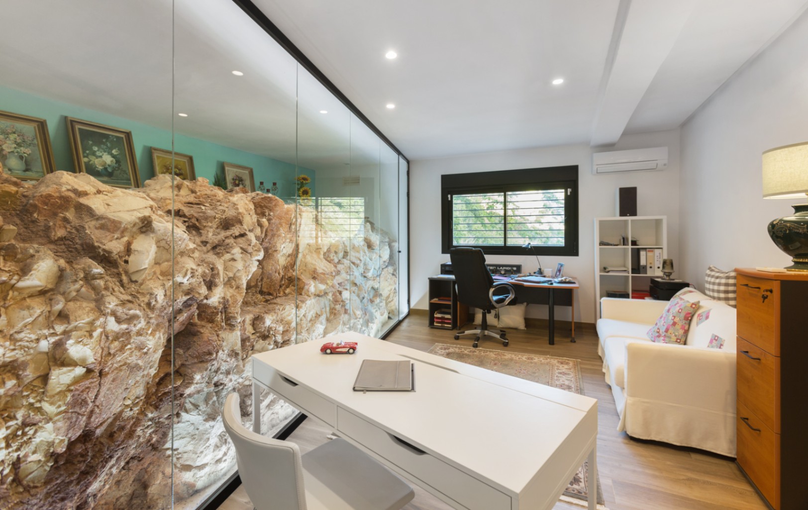 Modernes Arbeitszimmer mit einer Glaswand, die eine natürliche Felsformation integriert. Ausgestattet mit weißem Schreibtisch, Sofa und Einbaustrahlern.