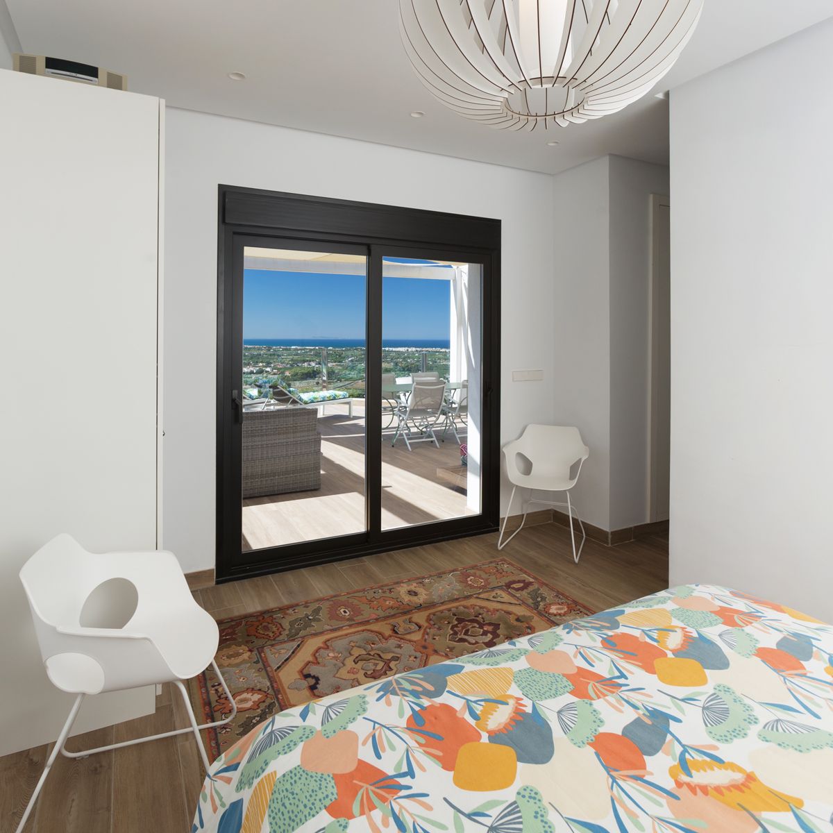 Modernes Schlafzimmer mit Glasschiebetür zu einer möblierten Terrasse mit Meerblick. Ausgestattet mit einer skulpturalen Pendelleuchte und weißen Designstühlen.