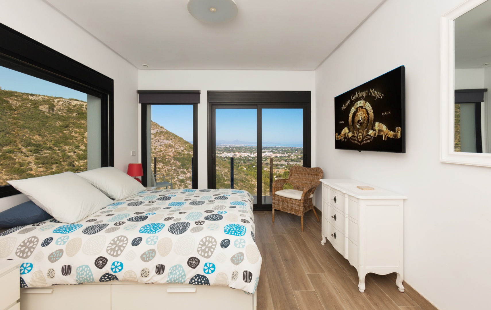 Schlafzimmer mit Panoramablick auf Berge und Meer, großen schwarz gerahmten Fenstern, Boden in Holzoptik, weißer Kommode und wandmontiertem Fernseher.