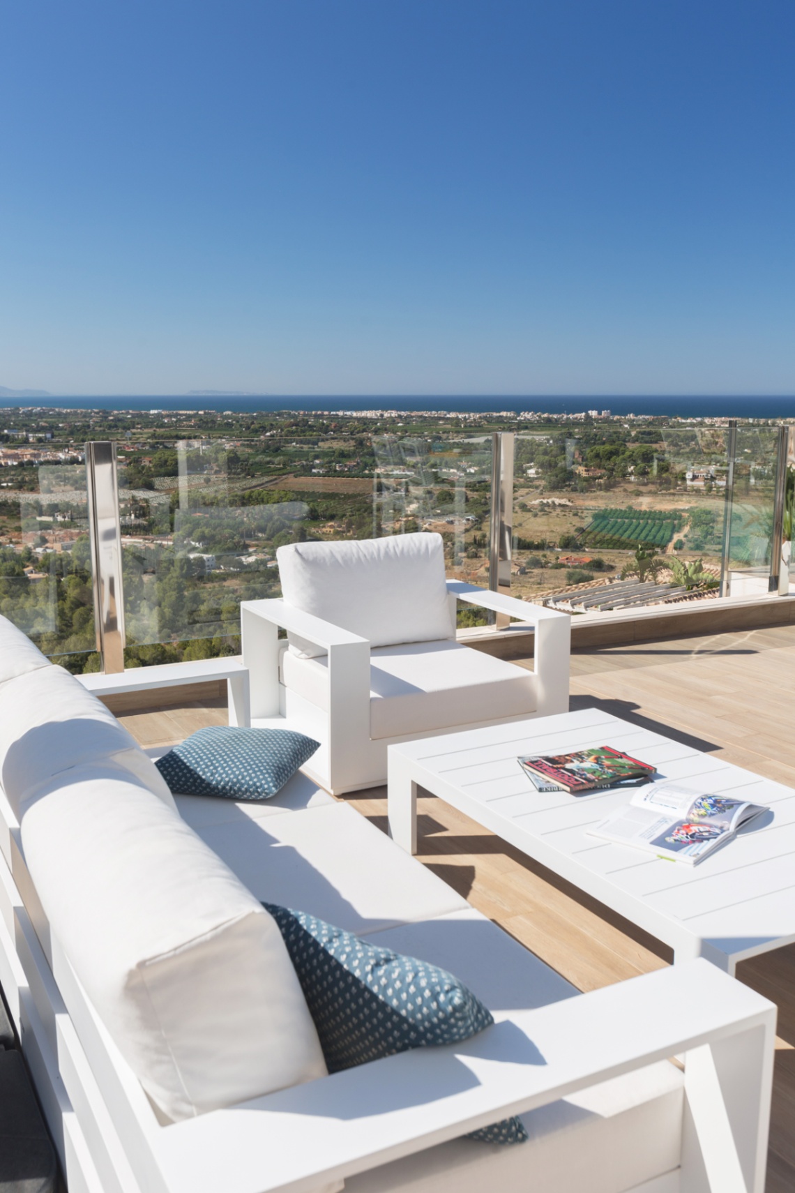 Terrasse mit modernen weißen Loungemöbeln und Glasgeländer, die einen Panoramablick auf die Küstenlandschaft und das Mittelmeer bietet.