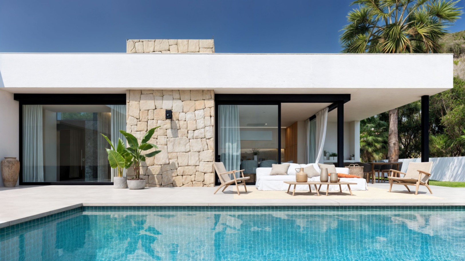 Außenansicht einer modernen Villa mit türkisfarbenem Pool, Natursteinmauerwerk und einem Loungebereich auf der großzügigen Fliesenterrasse.