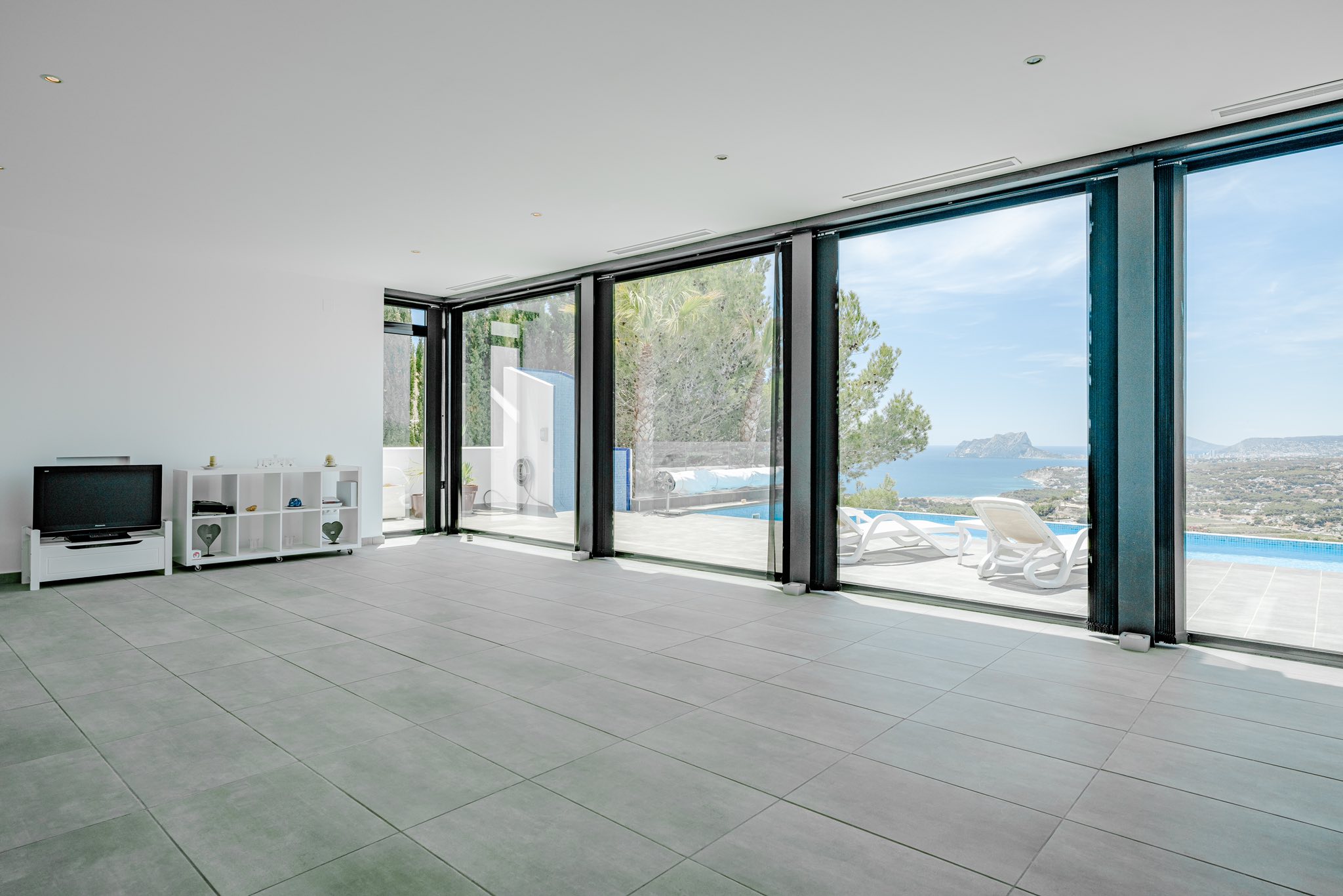 Großzügiger Wohnraum mit grauen Bodenfliesen und bodentiefen Glasfronten, die auf die Poolterrasse mit Panoramablick auf das Meer und den Peñón de Ifach führen.