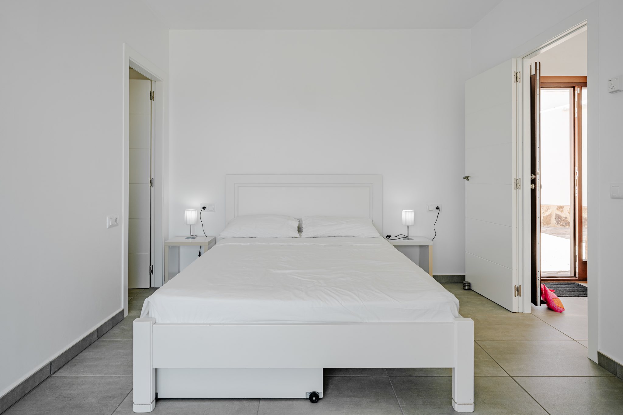 Minimalistisches Schlafzimmer mit weißem Holzbett, passenden Nachttischen und grauem Fliesenboden. Offene Türen führen zu angrenzenden Räumen und nach draußen.