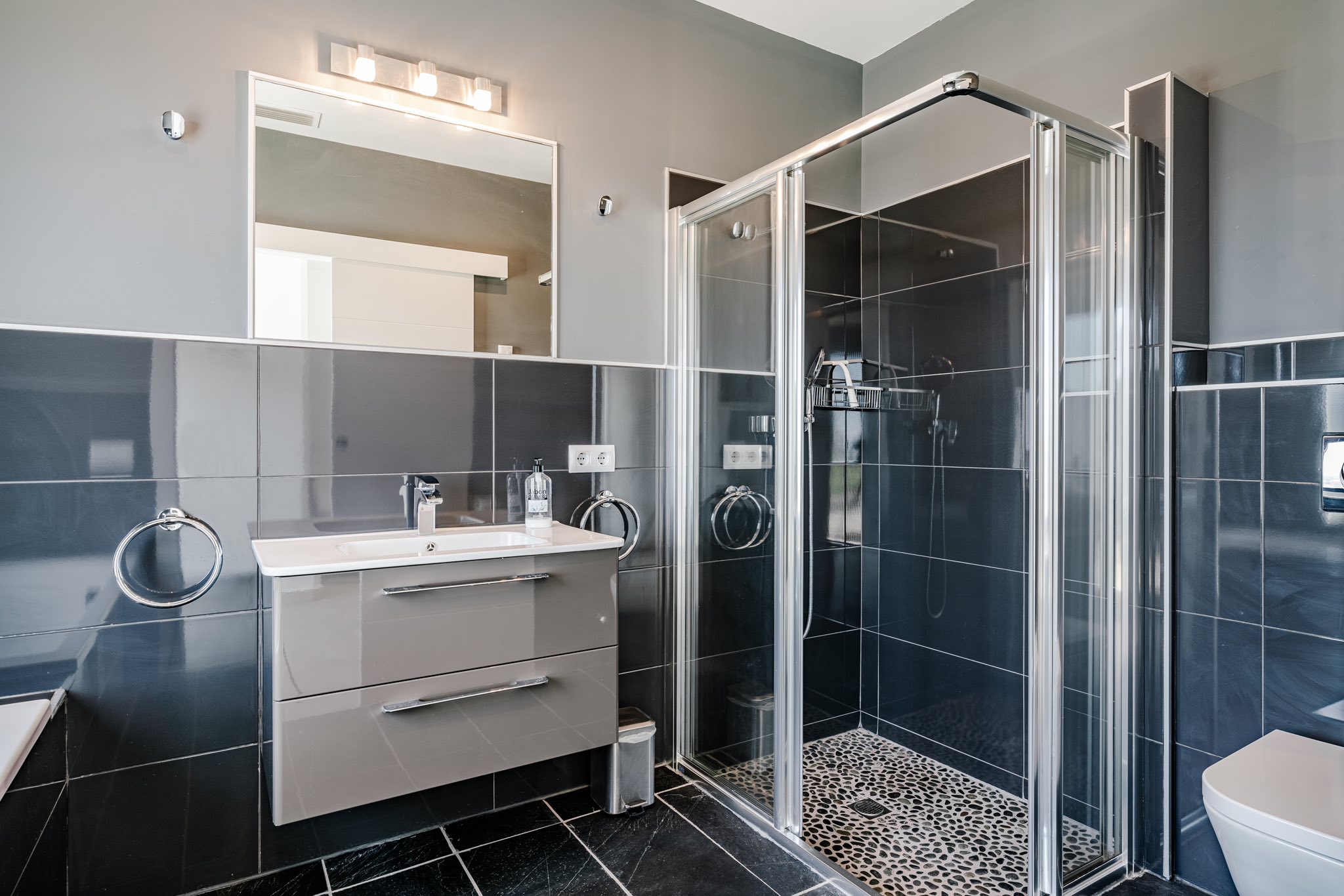 Modernes Badezimmer mit anthrazitfarbenen Glanzfliesen, schwebendem Waschtisch und einer Glasdusche mit Bodenbelag in Kieselstein-Optik.