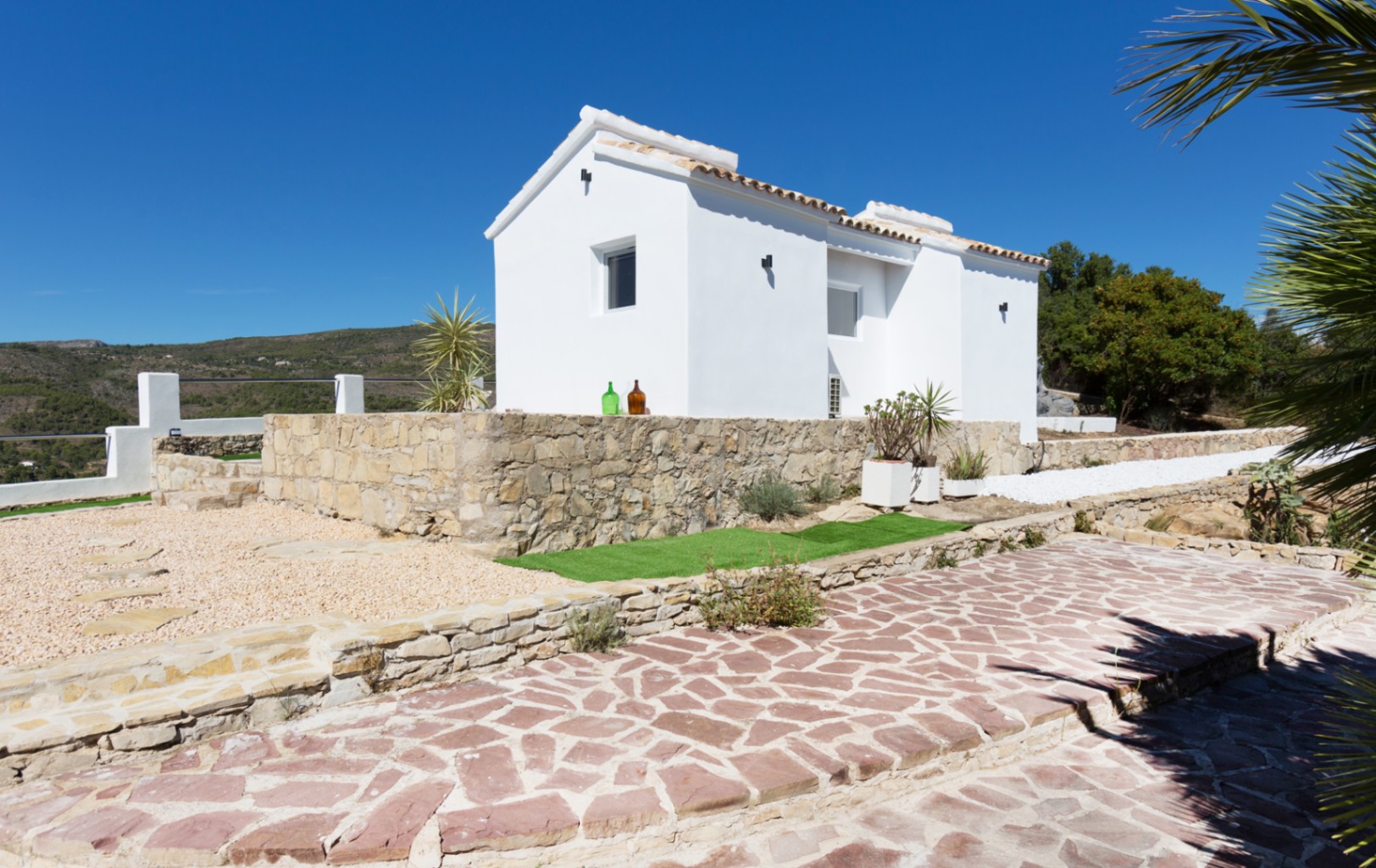 Außenansicht einer weiß getünchten Villa mit Natursteinmauern, traditionellem Ziegeldach und gepflastertem Weg vor einer bergigen Landschaftskulisse.