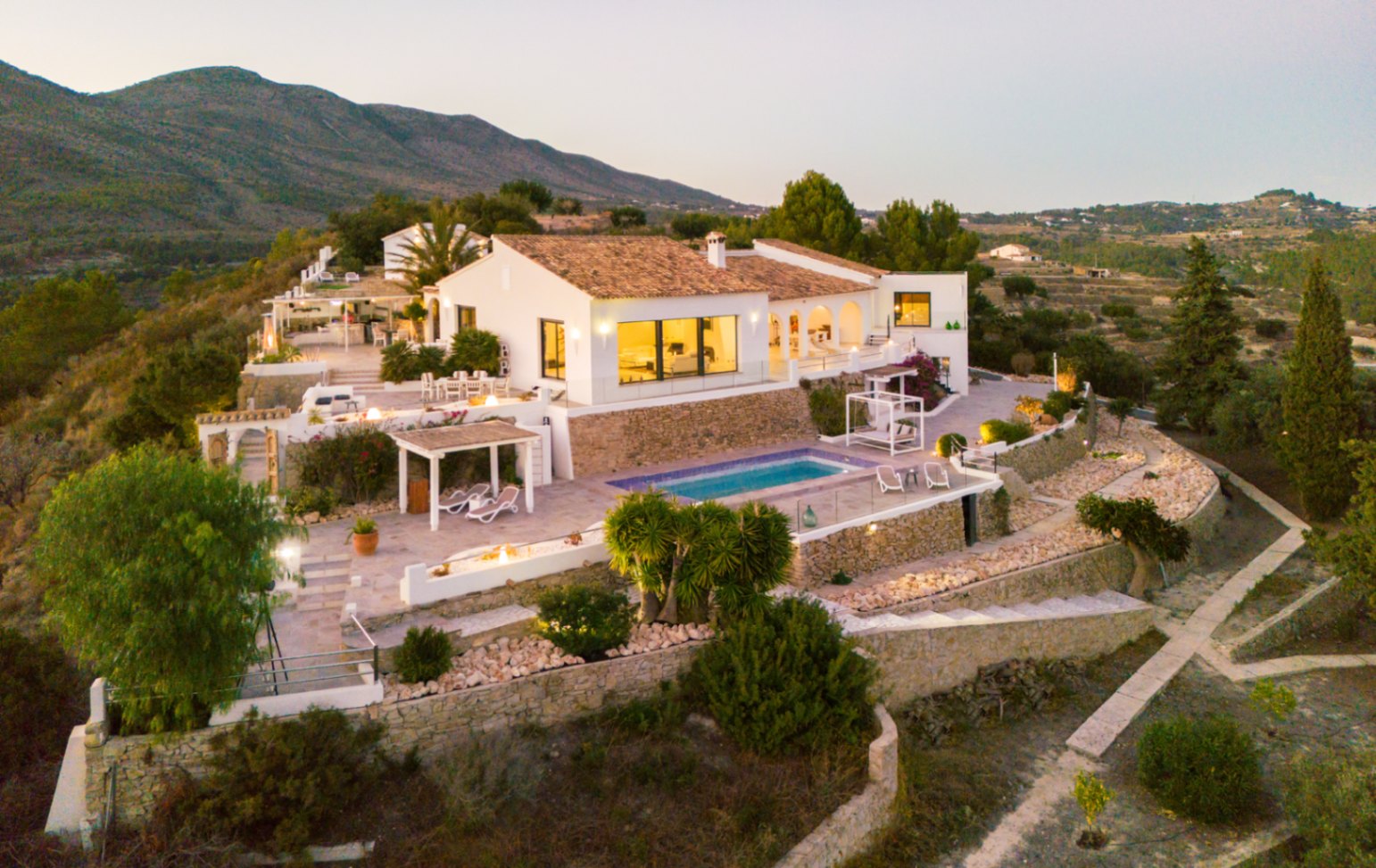 Erhöhte Ansicht einer mediterranen Villa mit weißen Fassaden, Terrakottadächern und abgestuften Terrassen mit Pool und Natursteinmauern.