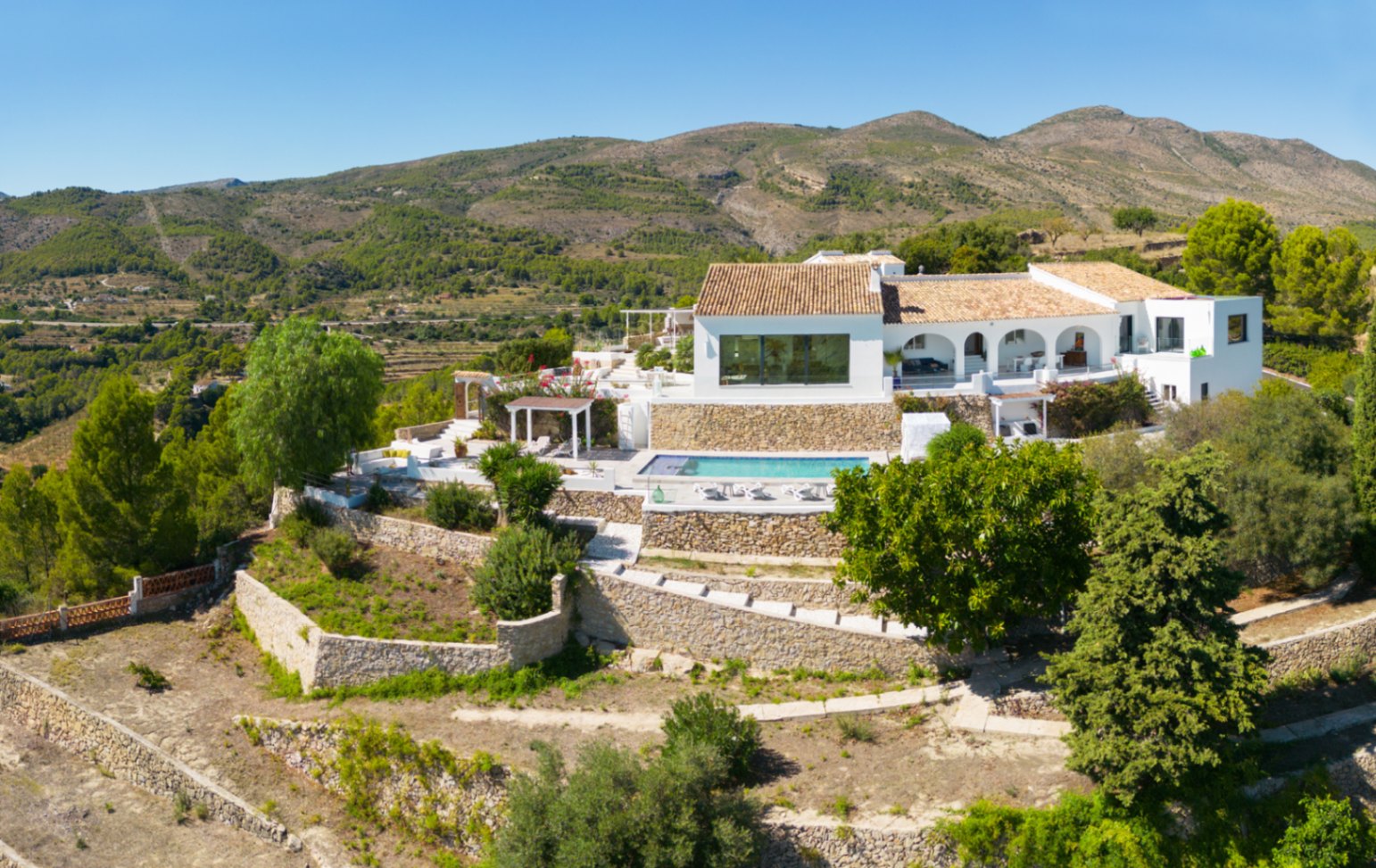 Erhöhte Ansicht einer weißen mediterranen Villa mit terrassierten Steingärten, privatem Pool und einer Rundbogen-Veranda vor einer bergigen Kulisse.