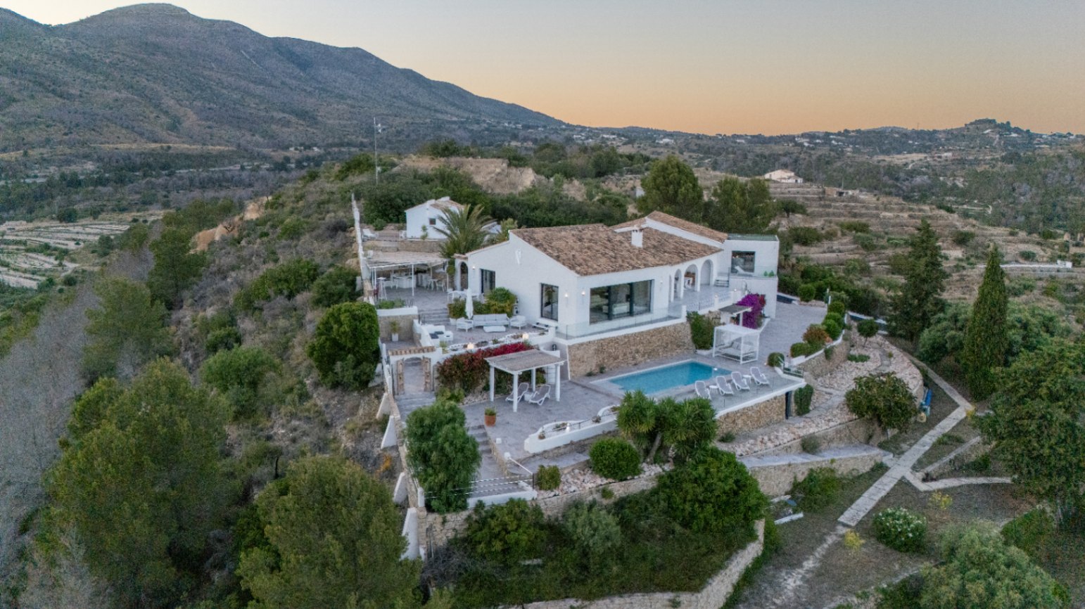 Erhöhte Ansicht einer weißen mediterranen Villa mit terrassierten Steinmauern, Pool und mehreren Außenbereichen vor einer bergigen Kulisse.