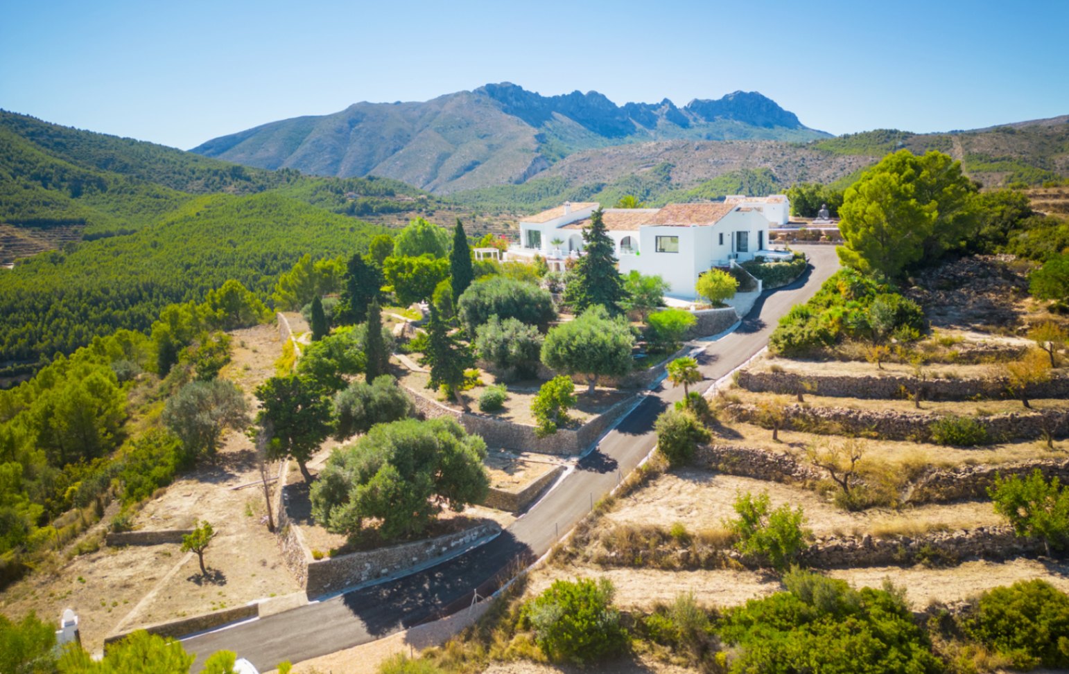 Luftaufnahme einer weißen Villa in Berglandschaft mit Steinterrassen, mediterranen Gärten und Panoramablick auf die umliegenden Pinienwälder.