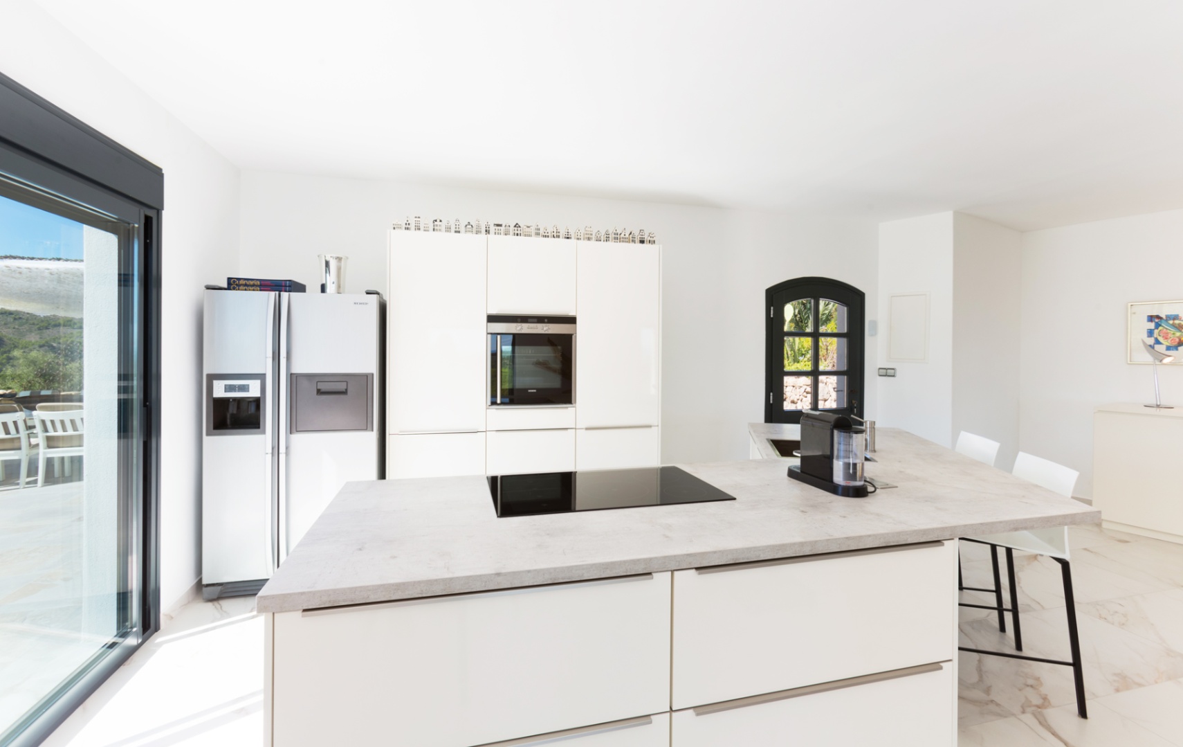 Moderne Küche mit Kochinsel in Steinoptik, Induktionsfeld, weißen Schränken und Side-by-Side-Kühlschrank. Marmorboden und viel Tageslicht.