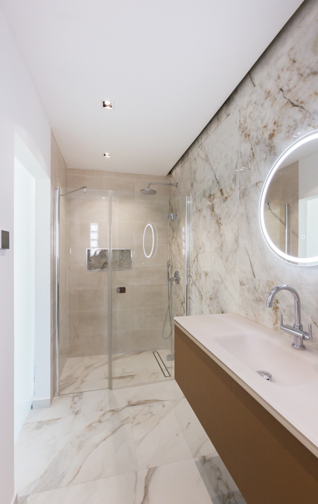 Modernes Badezimmer mit begehbarer Glasdusche, Wandfliesen in Marmoroptik, schwebendem Waschtisch und einem kreisförmigen Spiegel mit LED-Hintergrundbeleuchtung.