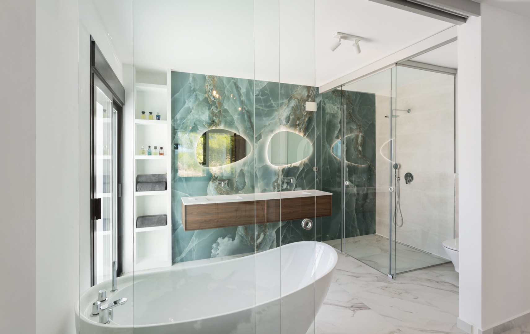 Modernes Badezimmer mit grüner Onyx-Wand, freistehender Badewanne und schwebendem Doppelwaschtisch aus Holz. Mit organisch geformten, hinterleuchteten Spiegeln und Glasdusche.