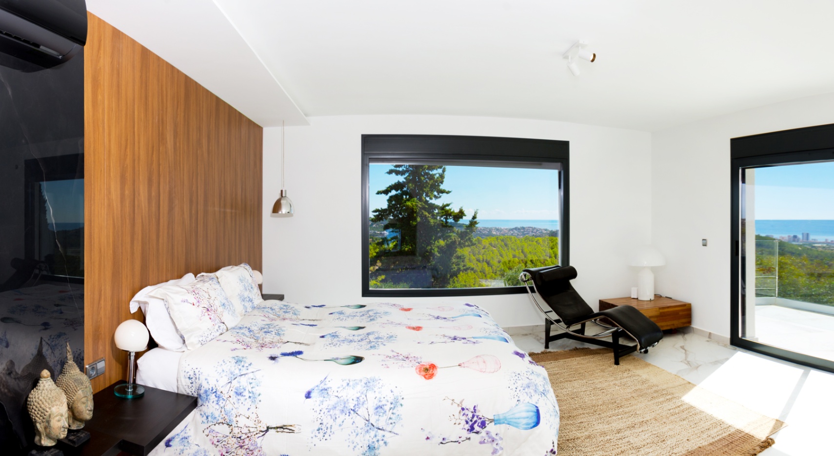 Modernes Schlafzimmer mit Akzentwand aus Holz, Marmorboden und großem Panoramafenster mit Blick auf das Meer und den Wald. Inklusive Designer-Chaiselongue.