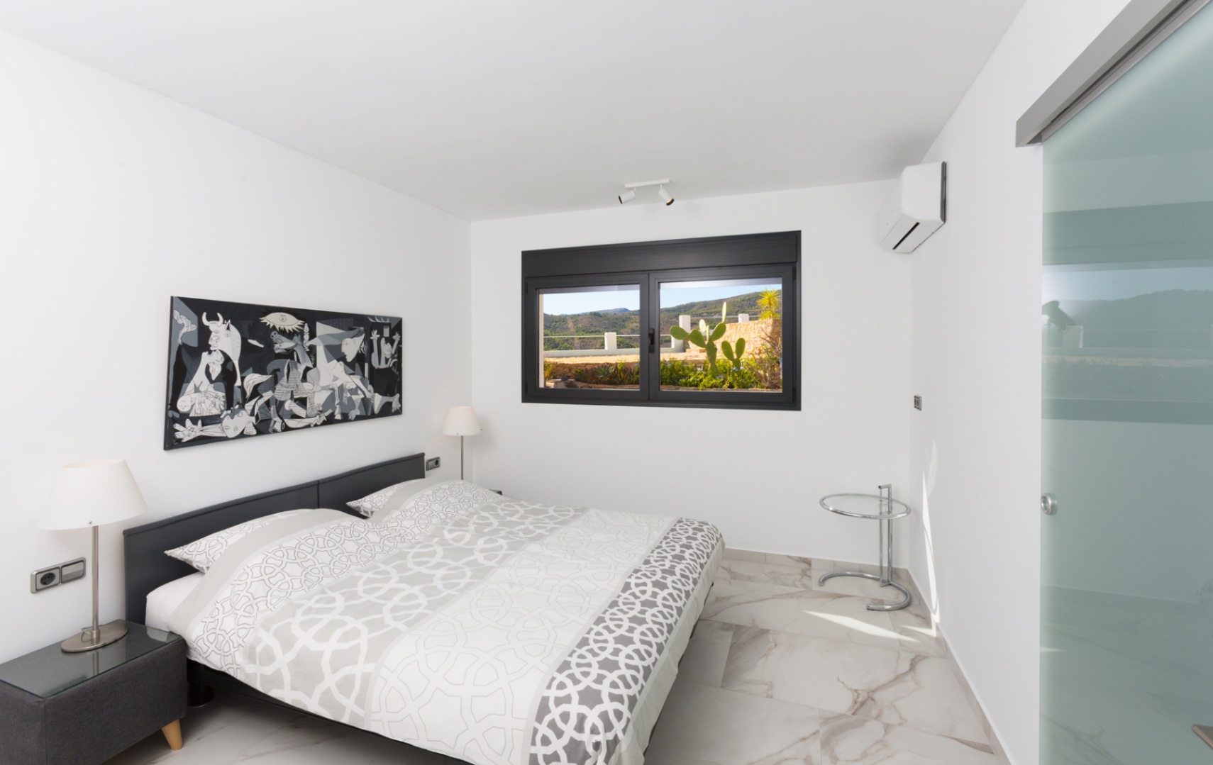 Modernes Schlafzimmer mit Boden in Marmoroptik, monochromem Kunstwerk und schwarzem Fensterrahmen mit Blick auf eine Terrasse mit Kakteen und Hügeln.