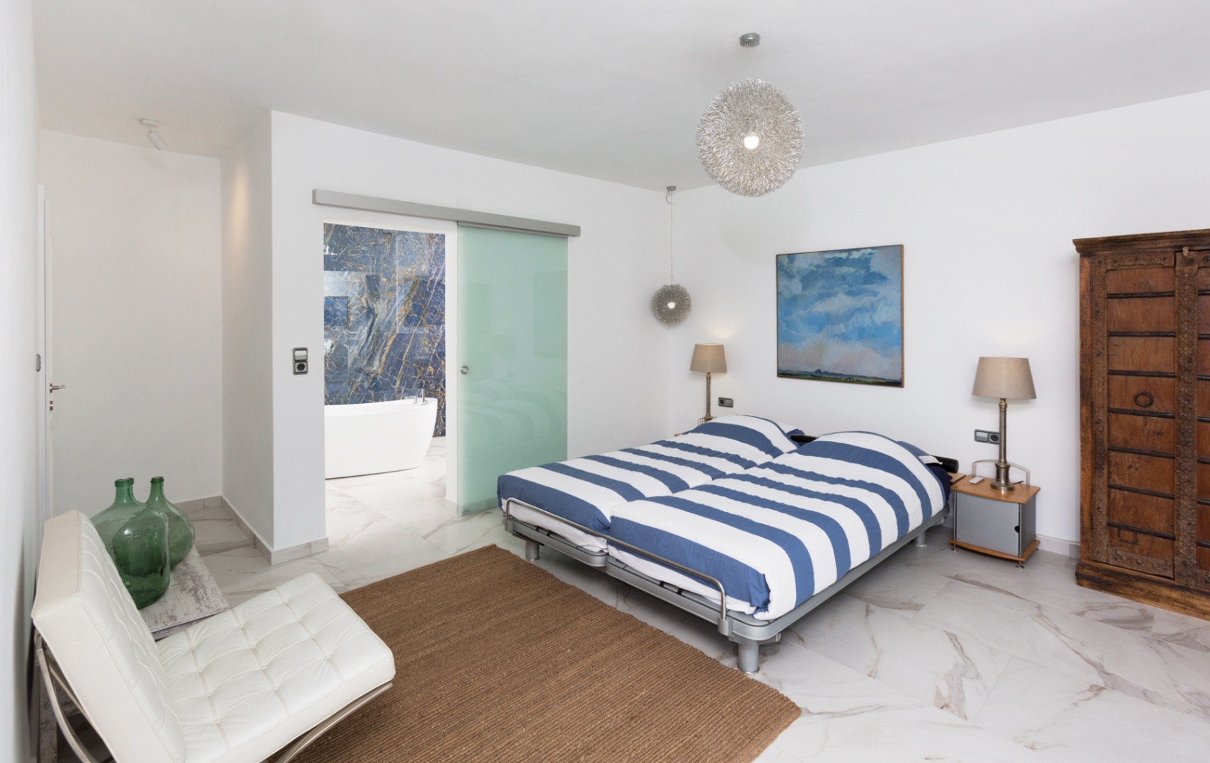 Modernes Schlafzimmer mit Marmorboden, blau gestreifter Bettwäsche und Glasschiebetür zum Bad mit freistehender Badewanne und blauer Natursteinwand.