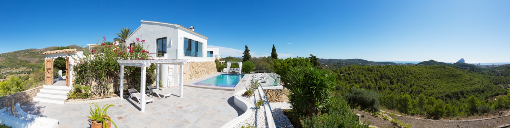 Panoramaterrasse mit Pool, weißer Pergola und Steinpflasterung, mit weitem Blick über ein bewaldetes Tal bis hin zum Mittelmeer und dem Peñón de Ifach.