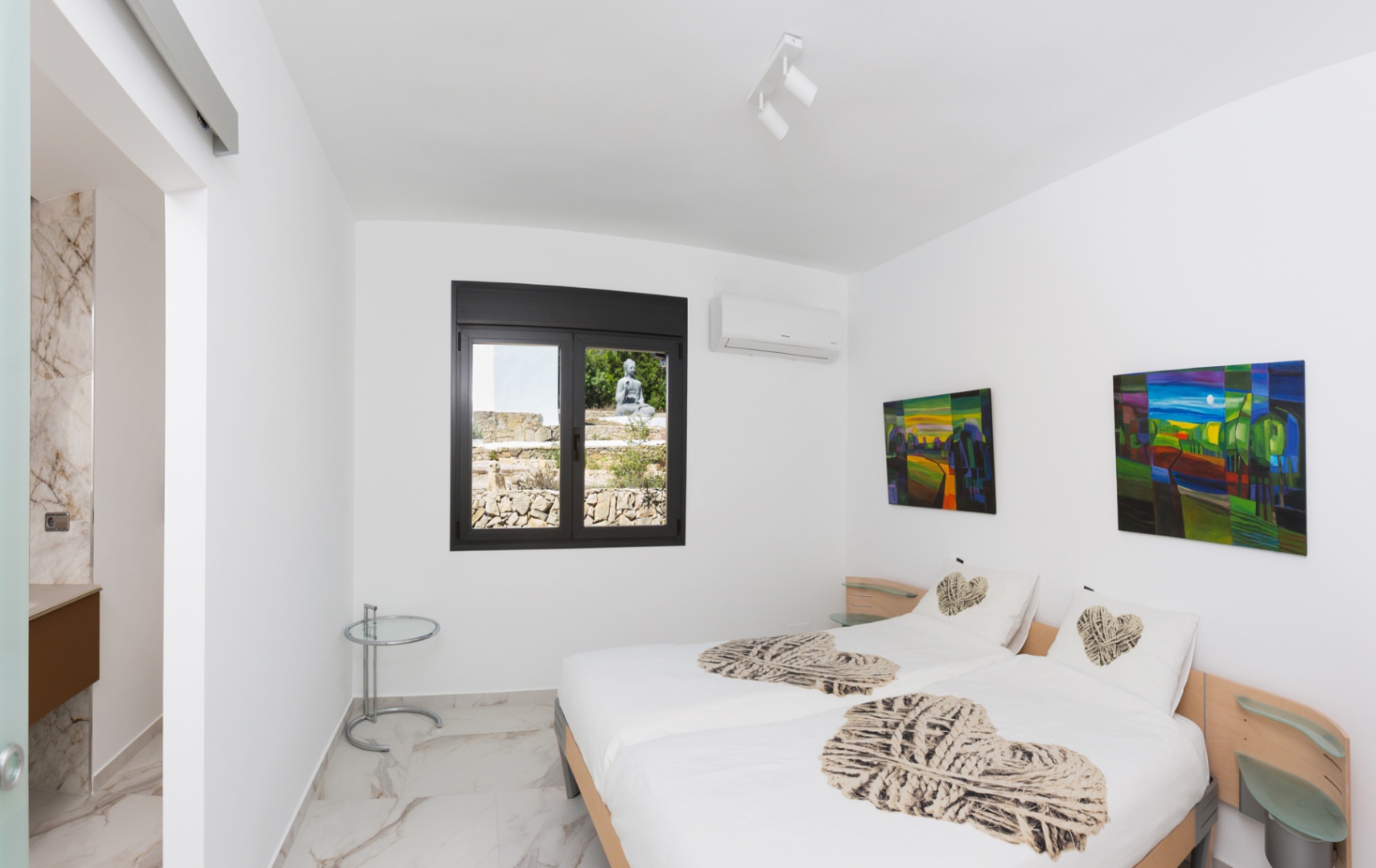 Schlafzimmer mit zwei Einzelbetten, Marmor-Optik-Fliesen und Fenster zum Steingarten. Ausgestattet mit Klimaanlage und direktem Zugang zum En-Suite-Bad.