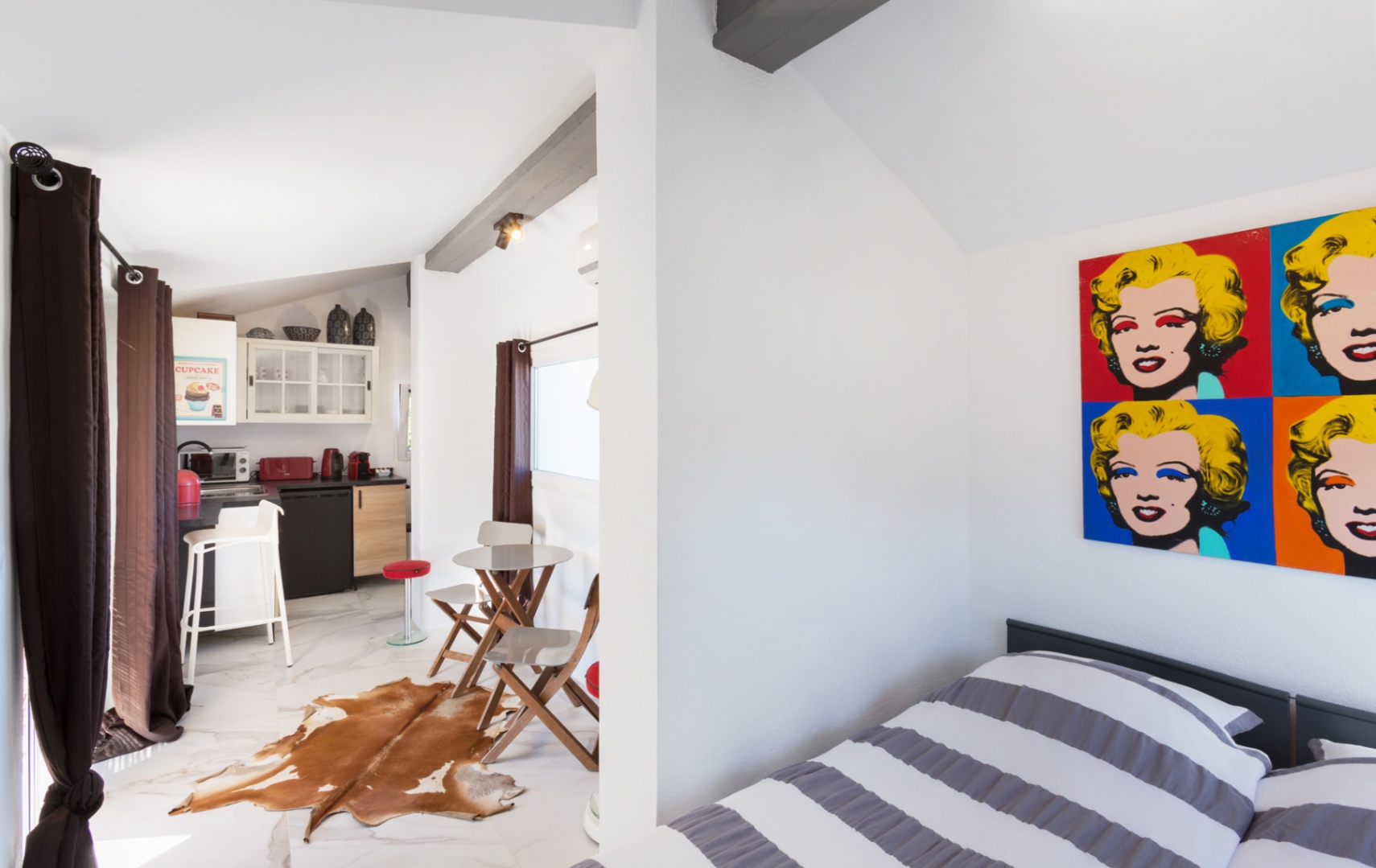 Studio mit Pop-Art-Dekor, dunklen Sichtbalken und Marmoroptik-Boden. Kompakte Küchenzeile mit roten Geräten und Essbereich mit echtem Kuhfellteppich.