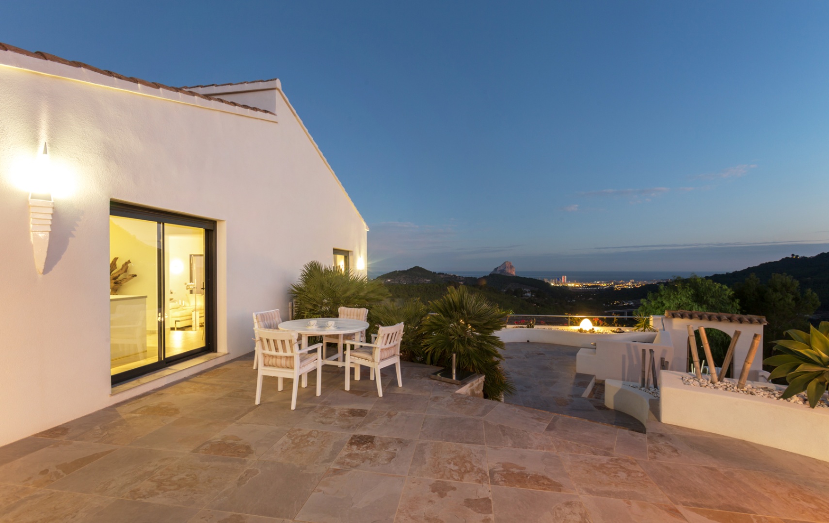 Terrasse mit Natursteinboden, weißer Sitzgruppe und Panoramablick auf die Küste von Calpe und den Peñón de Ifach in der Abenddämmerung.