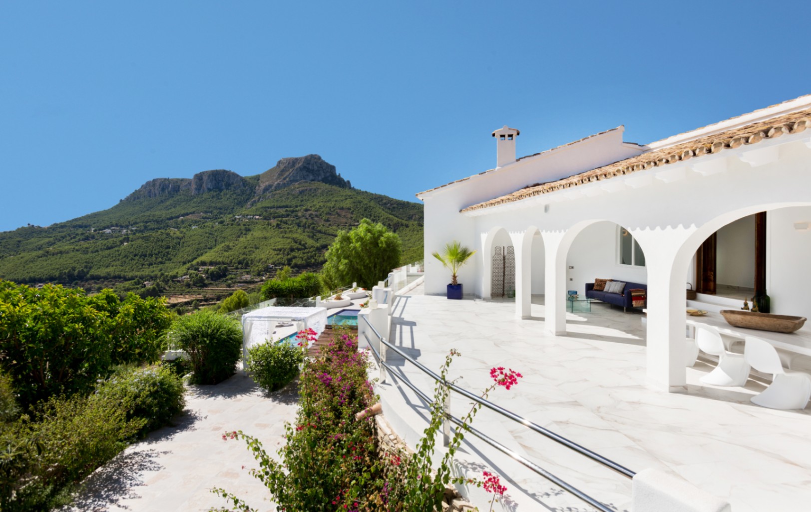 Weiße mediterrane Villa mit Bogenterrasse, Marmorboden, Außenlounge und weitem Blick auf die üppige Berglandschaft unter blauem Himmel.