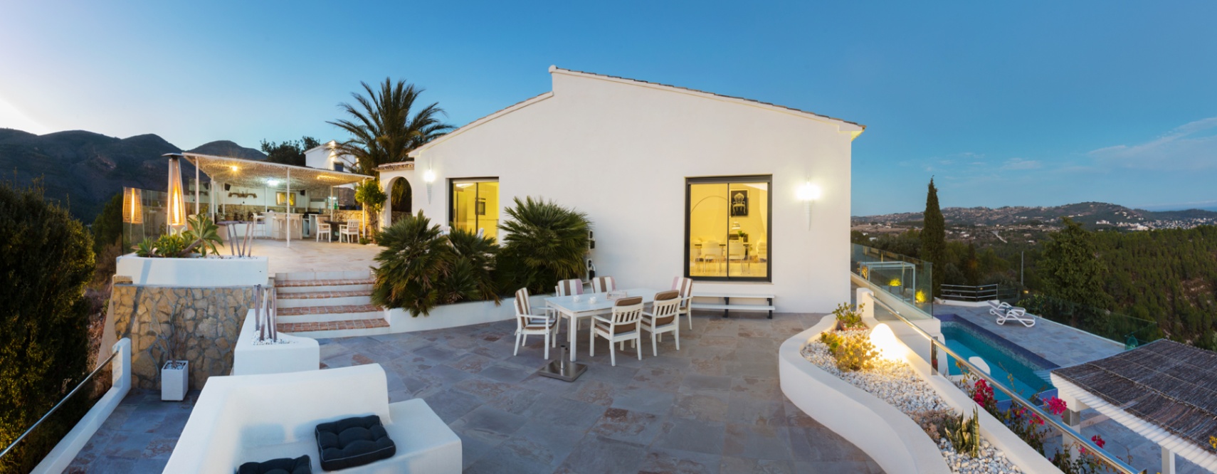 Weitläufige Steinterrasse in der Dämmerung mit Essbereich, integrierten Pflanzgefäßen und Panoramablick auf das Tal neben einer weißen mediterranen Villa.