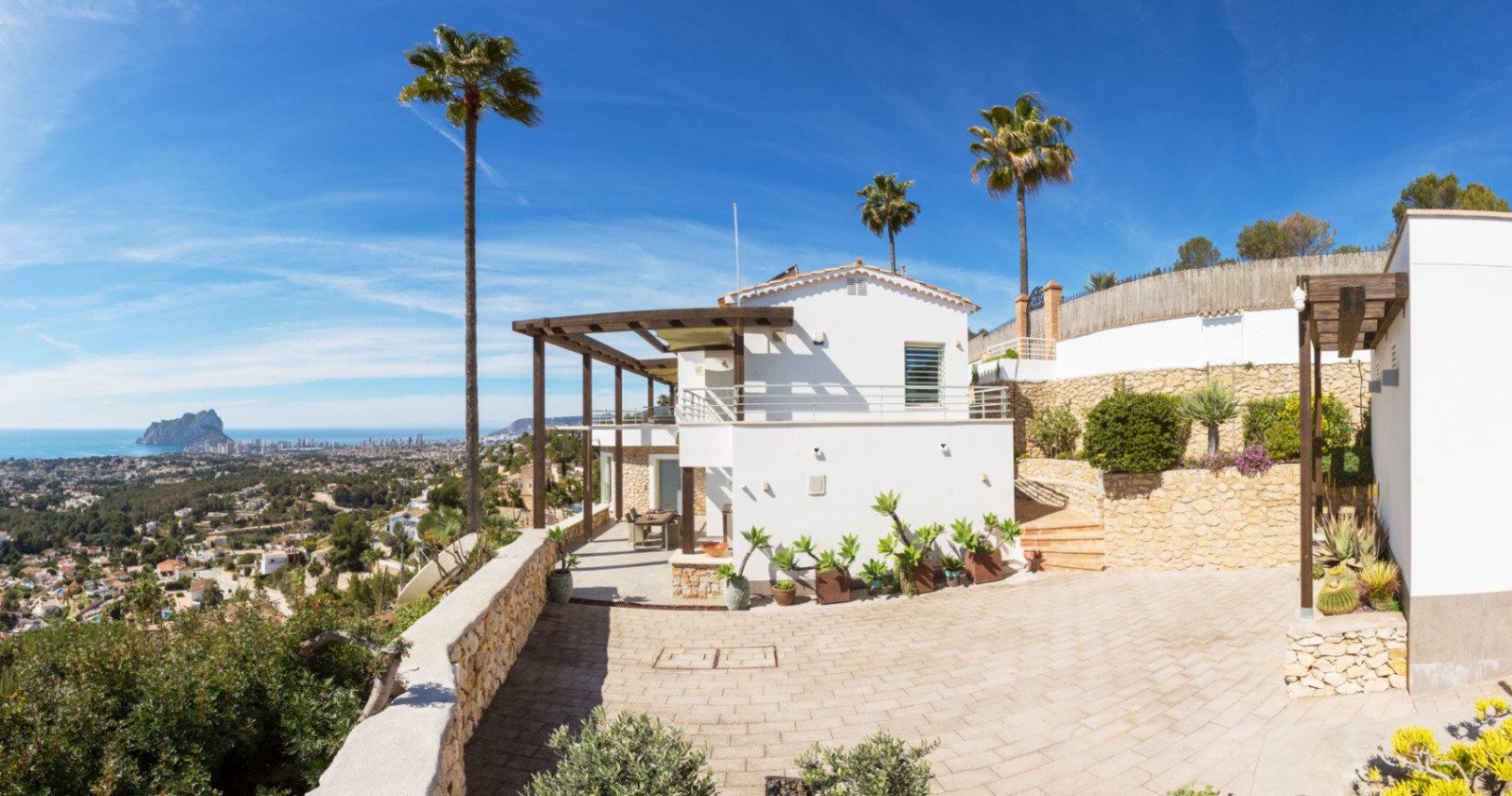 Außenansicht einer mediterranen Villa mit weißen Fassaden, Holzpergolen und Steinmauern, mit Panoramablick auf die Küste und den Peñón de Ifach.