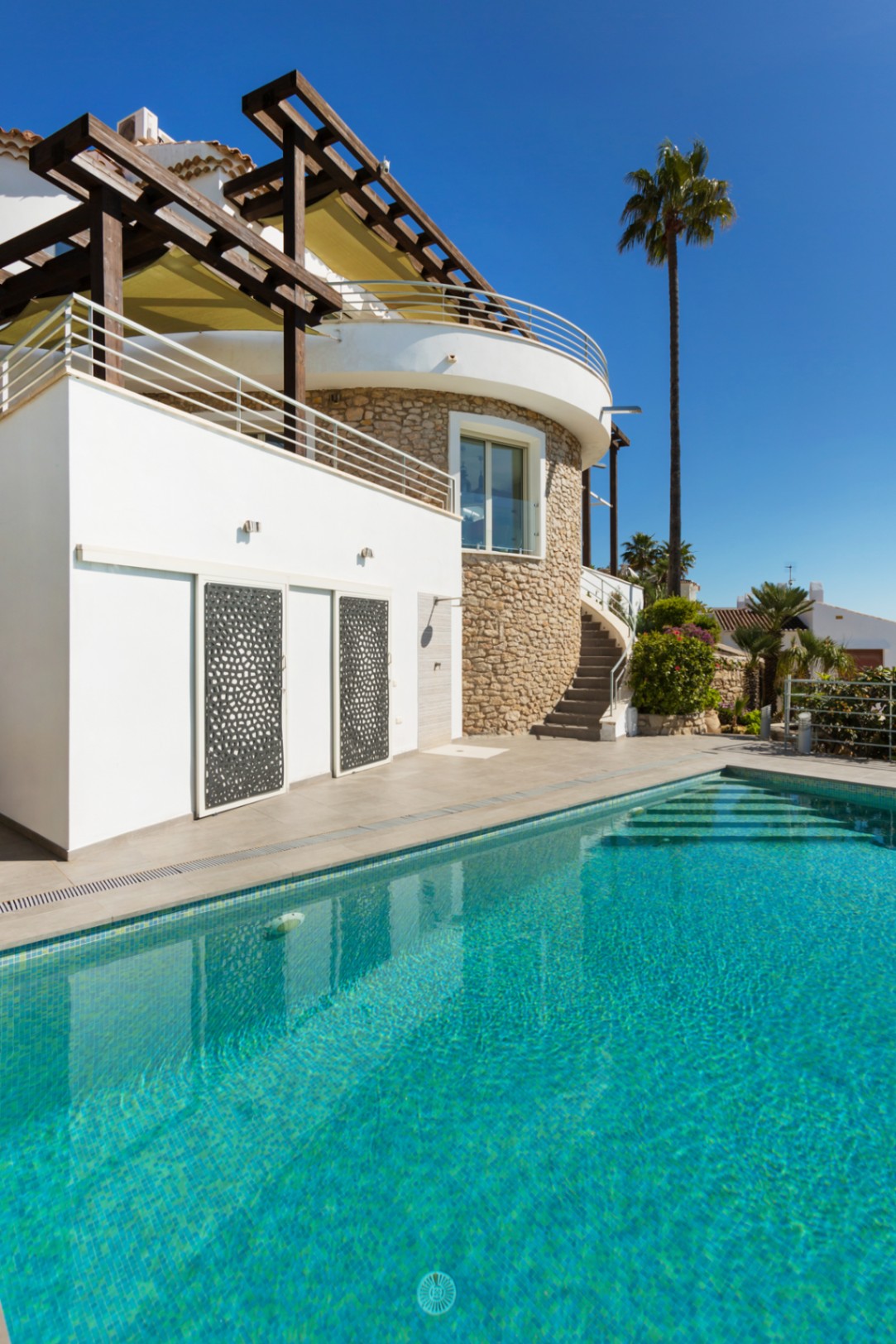 Außenansicht eines türkisfarbenen Pools neben einer weißen Villa mit runder Natursteinwand, Holzpergola und dekorativen Schiebeelementen aus Metall.